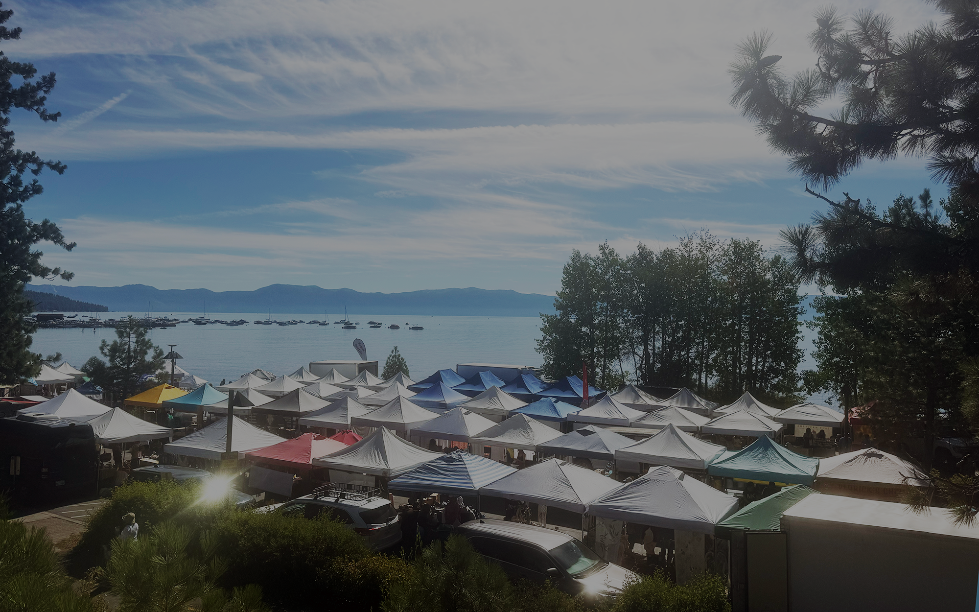 Farmers market tents fill Commons Beach beside Lake Tahoe, viewed from above.