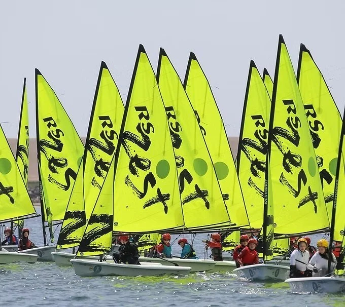 Bright RS Zest dinghies with young sailors gathered on the water during a youth sailing event.