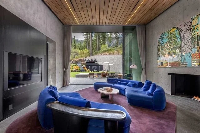 Living room with blue sectional sofa opening through a glass wall to an outdoor patio and forest view.