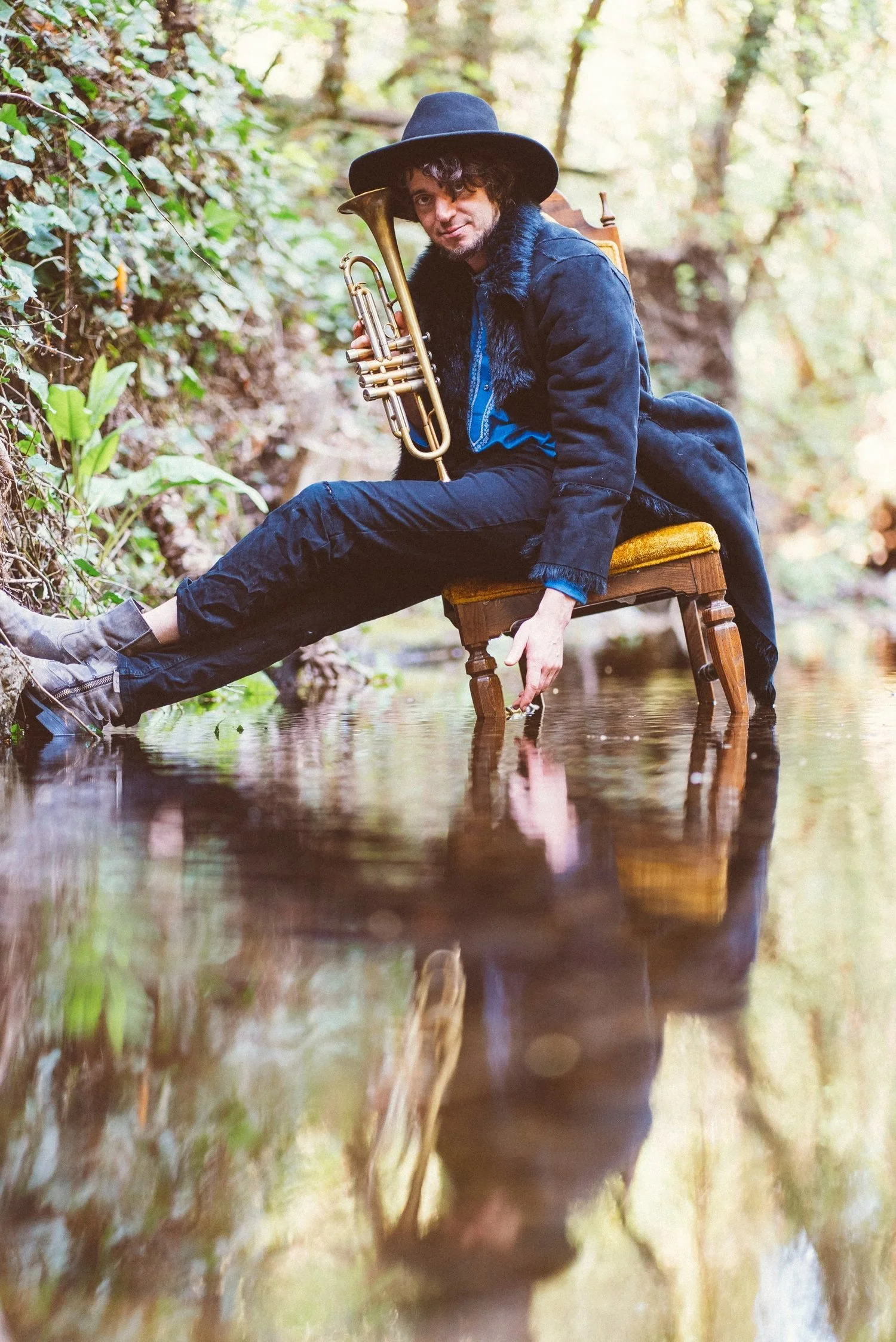 Balkan Bump holds a trumpet while seated on a wooden chair in shallow water surrounded by greenery.
