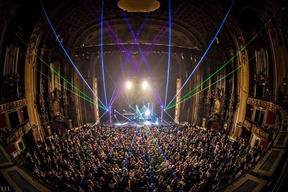 A packed theater faces a stage beneath multicolored concert lighting and laser beams.