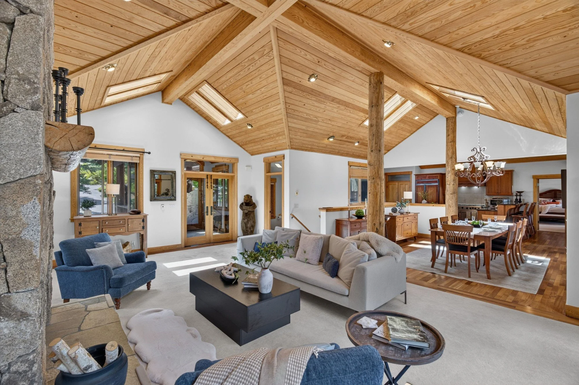 Great room with a vaulted wood ceiling, skylights, log posts and a stone fireplace, open to the dining area and kitchen