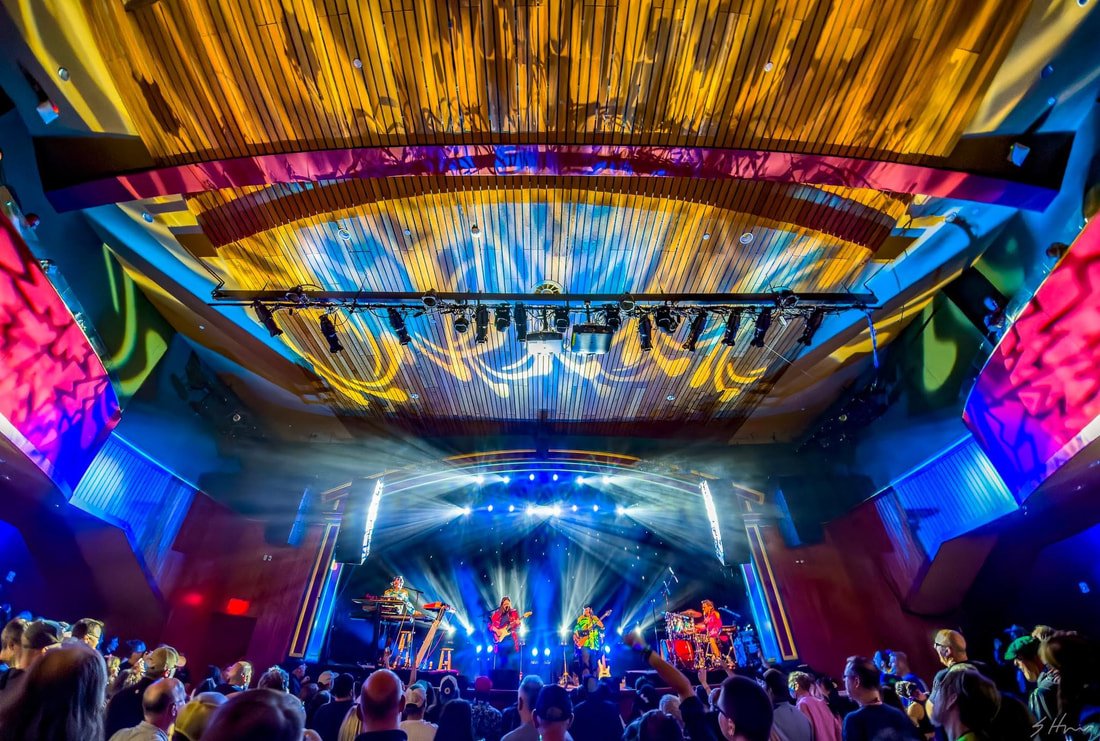 A concert crowd faces a brightly lit stage beneath a colorful patterned theater ceiling.