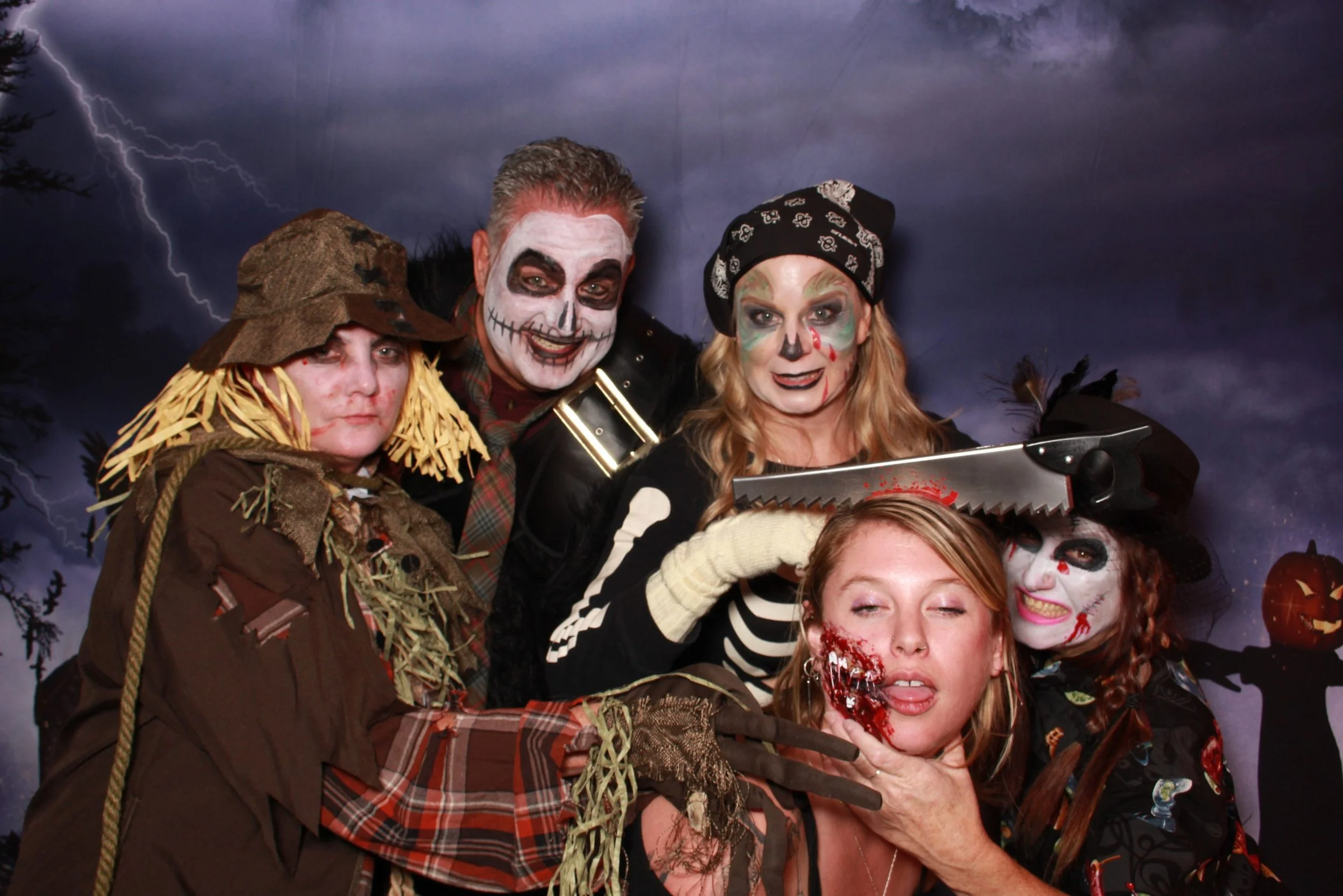 Five people in Halloween costumes and theatrical makeup pose with a prop saw against a stormy backdrop.