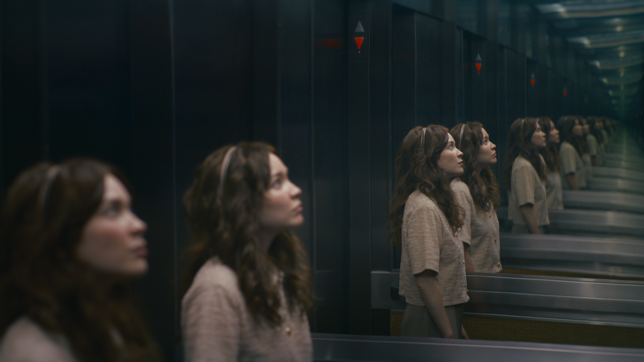 Movie still from Pocket Mirror. A young woman stands in an elevator, her mirror image repeating endlessly.
