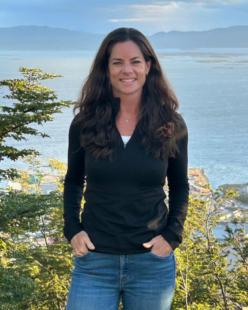 A woman with long brown hair smiling outdoors, standing in front of a scenic water view with mountains in the background, wearing a black zip-up jacket and blue jeans.