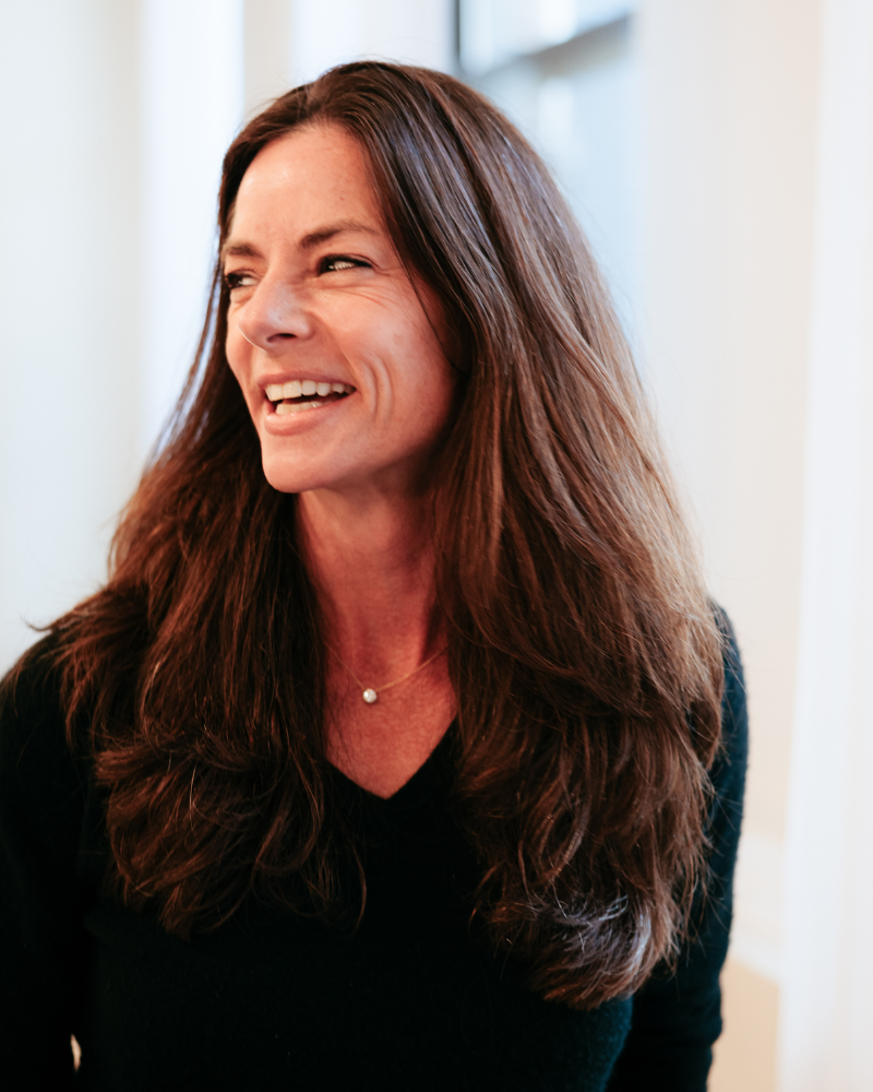 A woman with long, brown hair is smiling and looking to the side, wearing a black top and a pearl necklace, in a bright indoor setting.