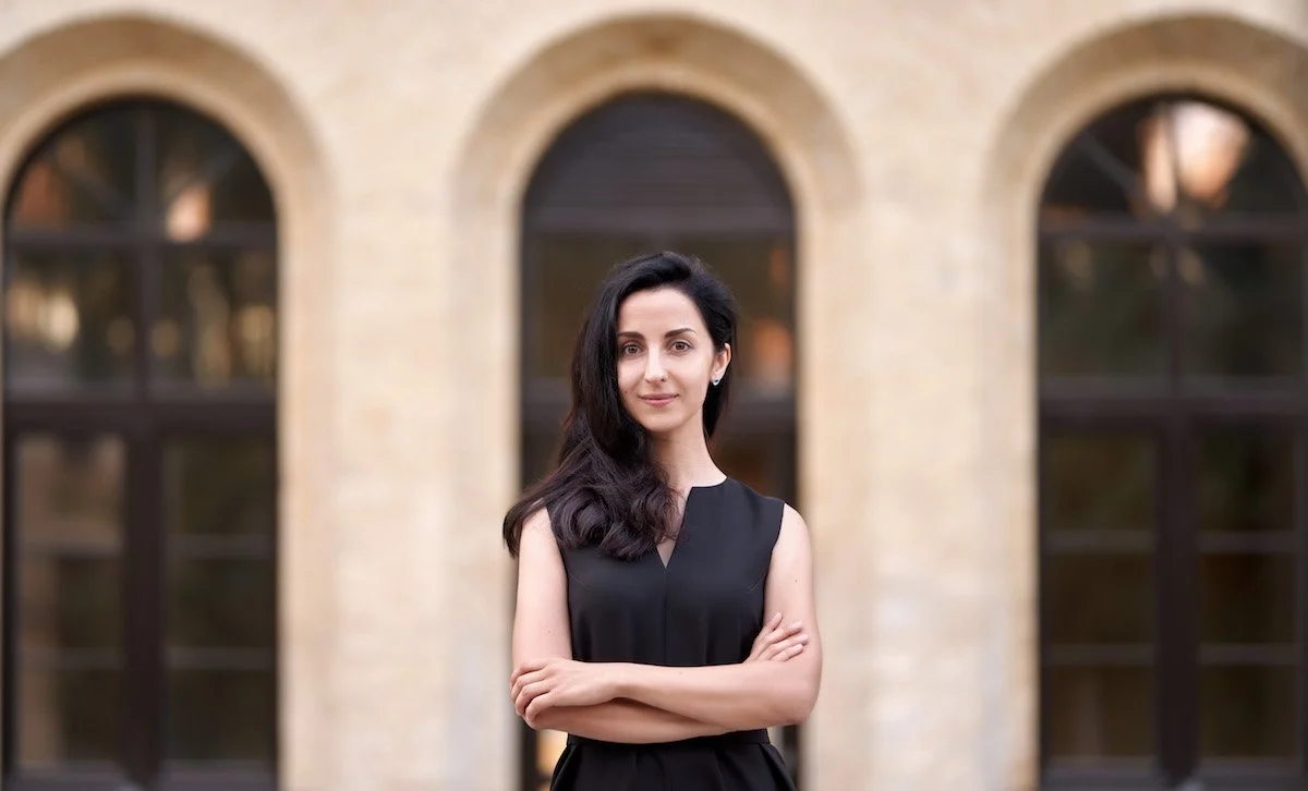 A woman with dark hair, wearing a sleeveless black dress, standing with arms crossed in front of a brick building with arched windows.