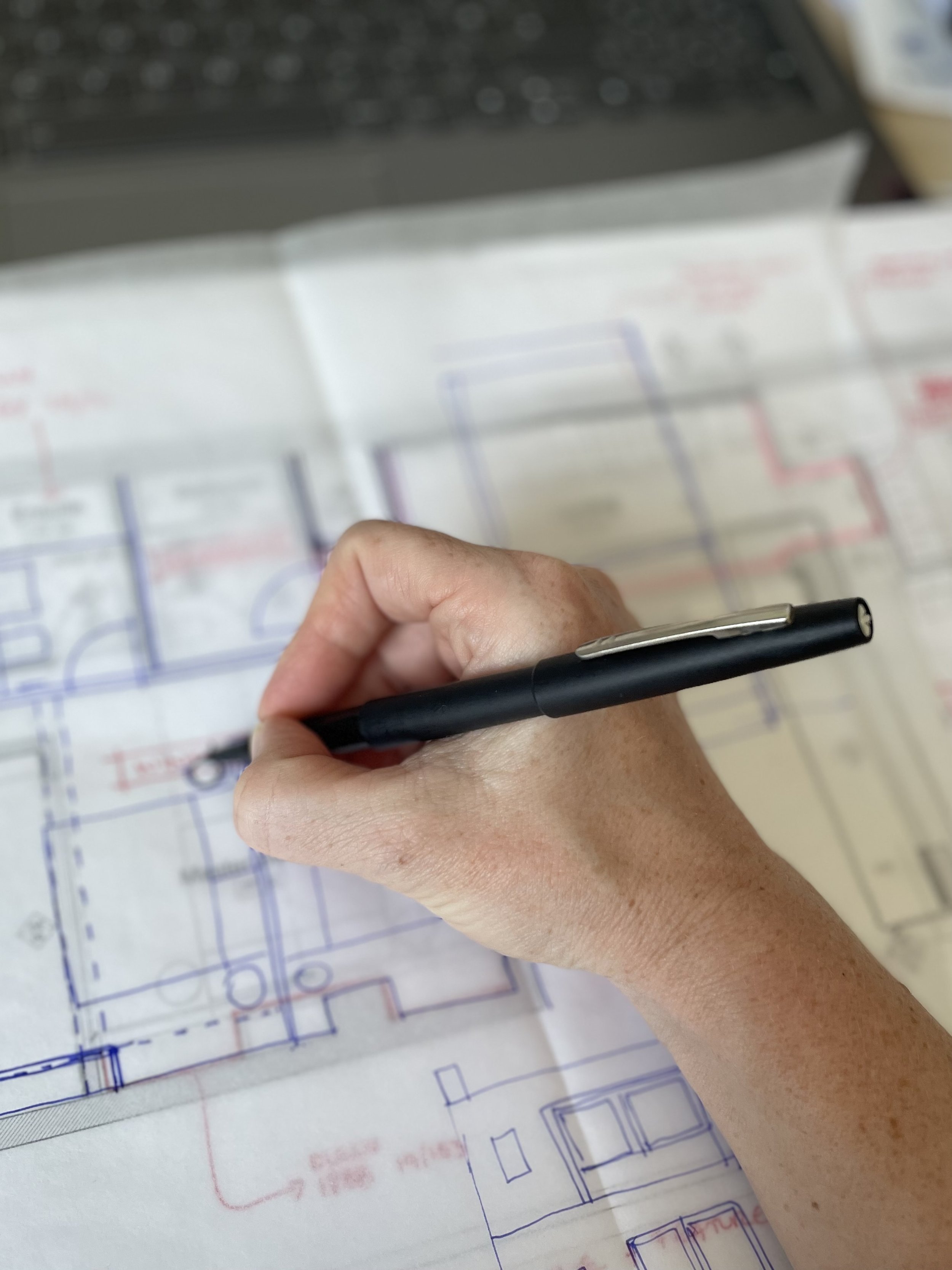 A person's hand holding a black pen, reviewing architectural blueprints on a table. The blueprints display detailed floor plans with red and blue ink. A laptop is partially visible in the background.