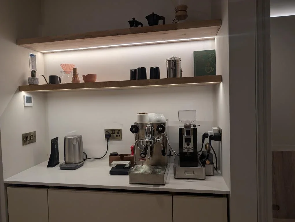 Coffee station with espresso machine, grinder, kettle, and shelves with cups, teapots, and decorative items.