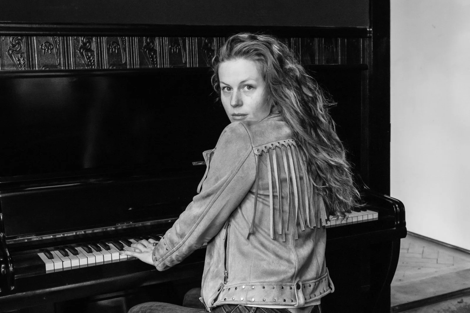 Schwarz-weiß-Fotografie einer jungen Frau mit langen, lockigen Haaren, die an einem Klavier sitzt und in die Kamera schaut