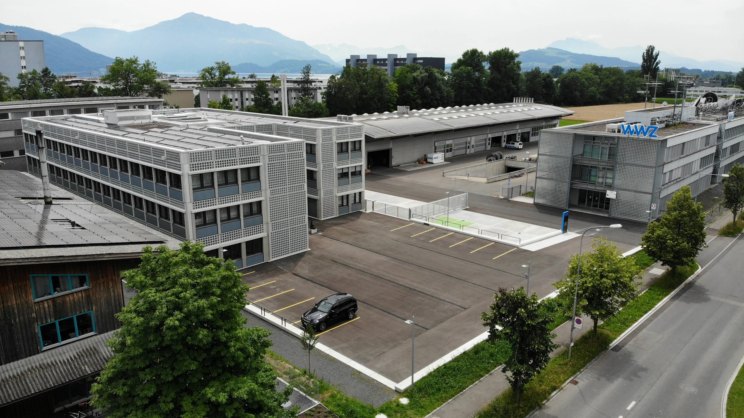 Drohnenaufnahme eines modernen Industriegebäudes mit einem Parkplatz und Bäumen, umgeben von einer ruhigen Straße, mit Bergen im Hintergrund.