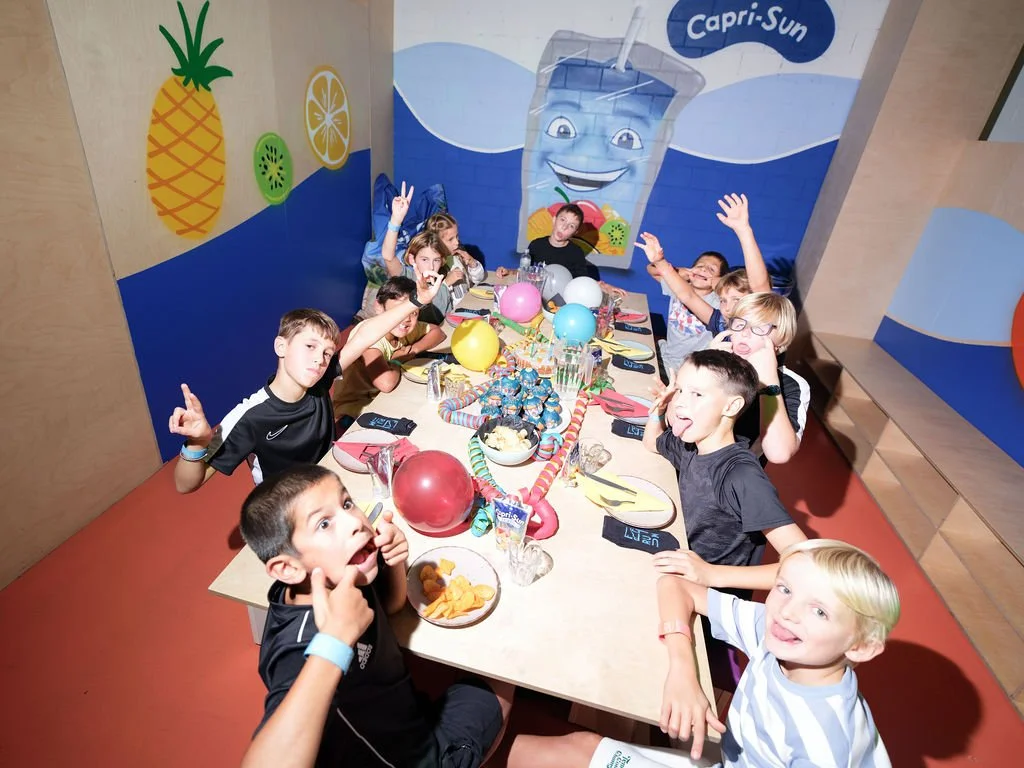 Kinder feiern ein Buffet-Abendessen bei einer Geburtstagsparty, mit bunten Luftballons, Kuchen und Partydekorationen, in einem Raum mit Wandbildern von Früchten und einem lachenden Glas.