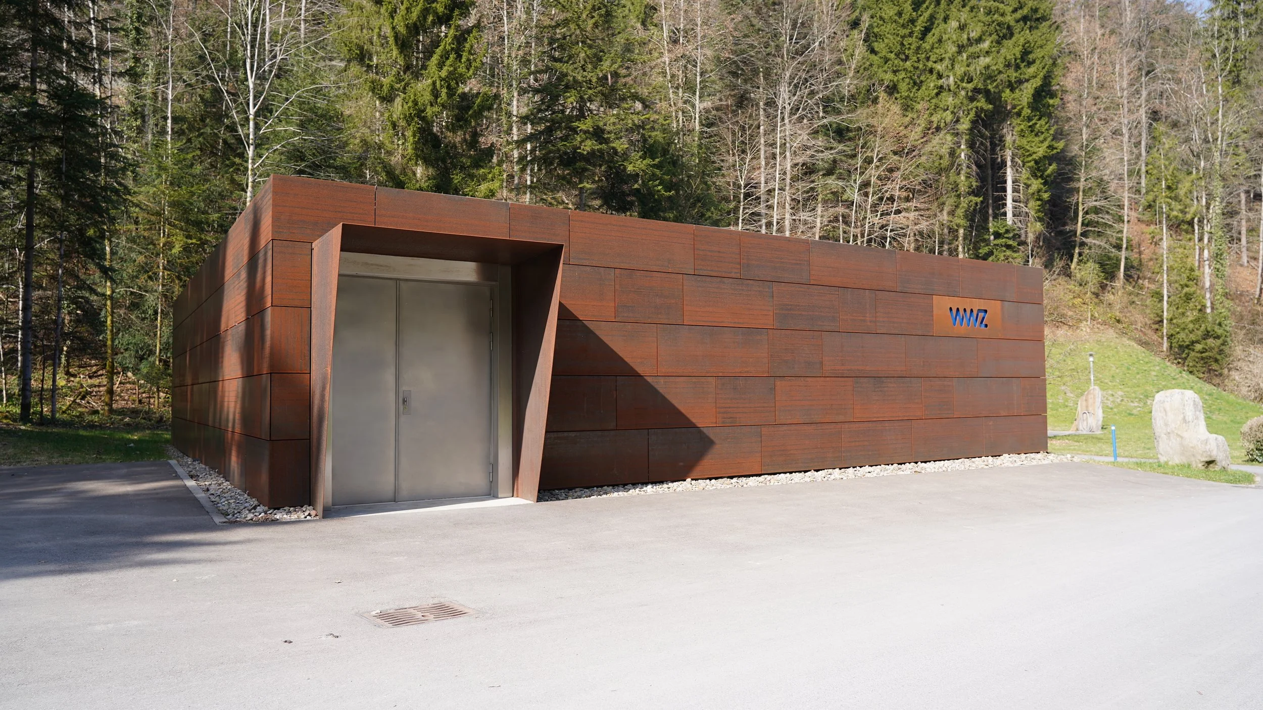 Moderner, rechteckiger Holzbau mit metallicem Eingang und blauer Aufschrift 'WWZ' an einer Straße inmitten eines Waldes.