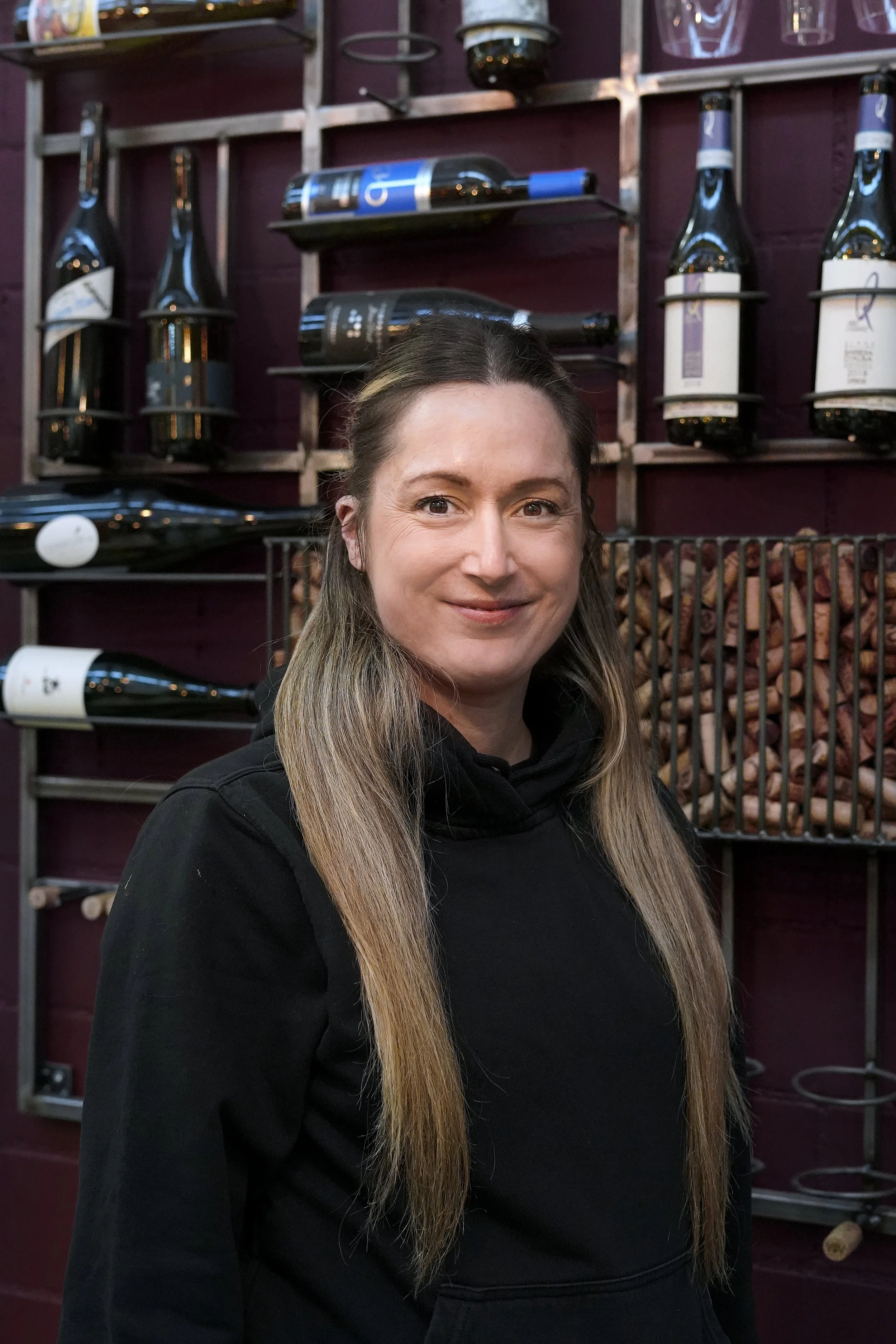Eine lachende Frau trägt eine schwarze Kleider und steht im Innenraum eines Restaurants oder cafes, mit schwarzen Holzregalen und Weinflaschen im Hintergrund.