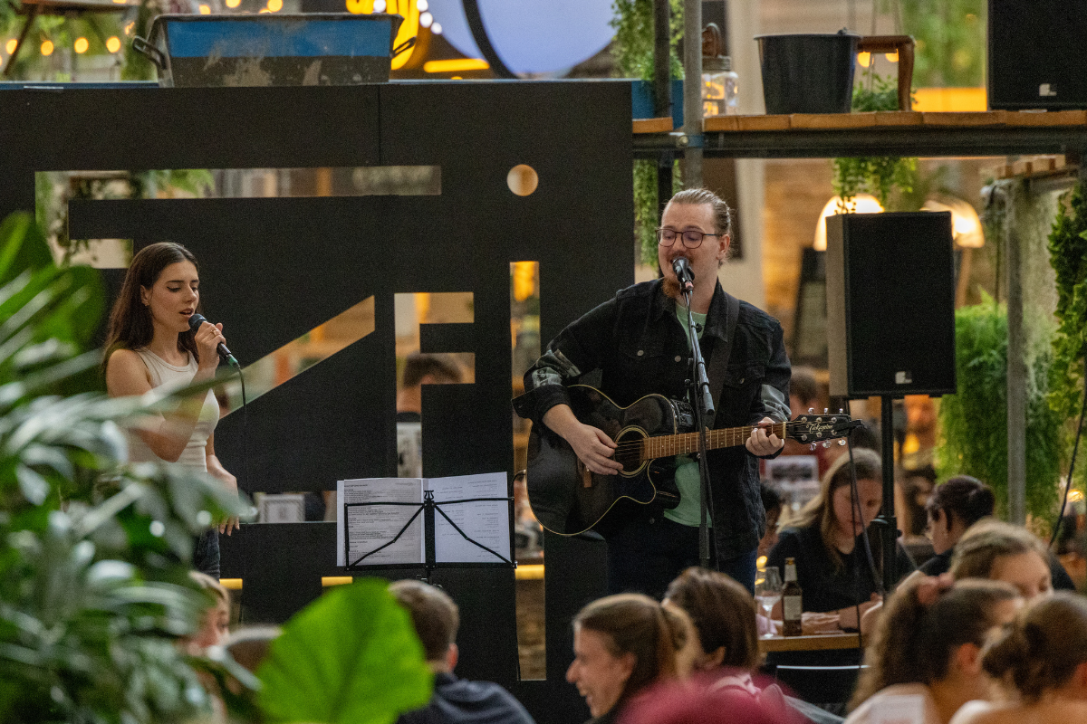 Zwei Musiker, eine Frau mit Microphone und eine Mann mit Gitarre, performen auf einer Bühne in der gemütlichen, gut besuchten Freiruum-Genusshalle, umgeben von grünen Pflanzen und live Publikum.