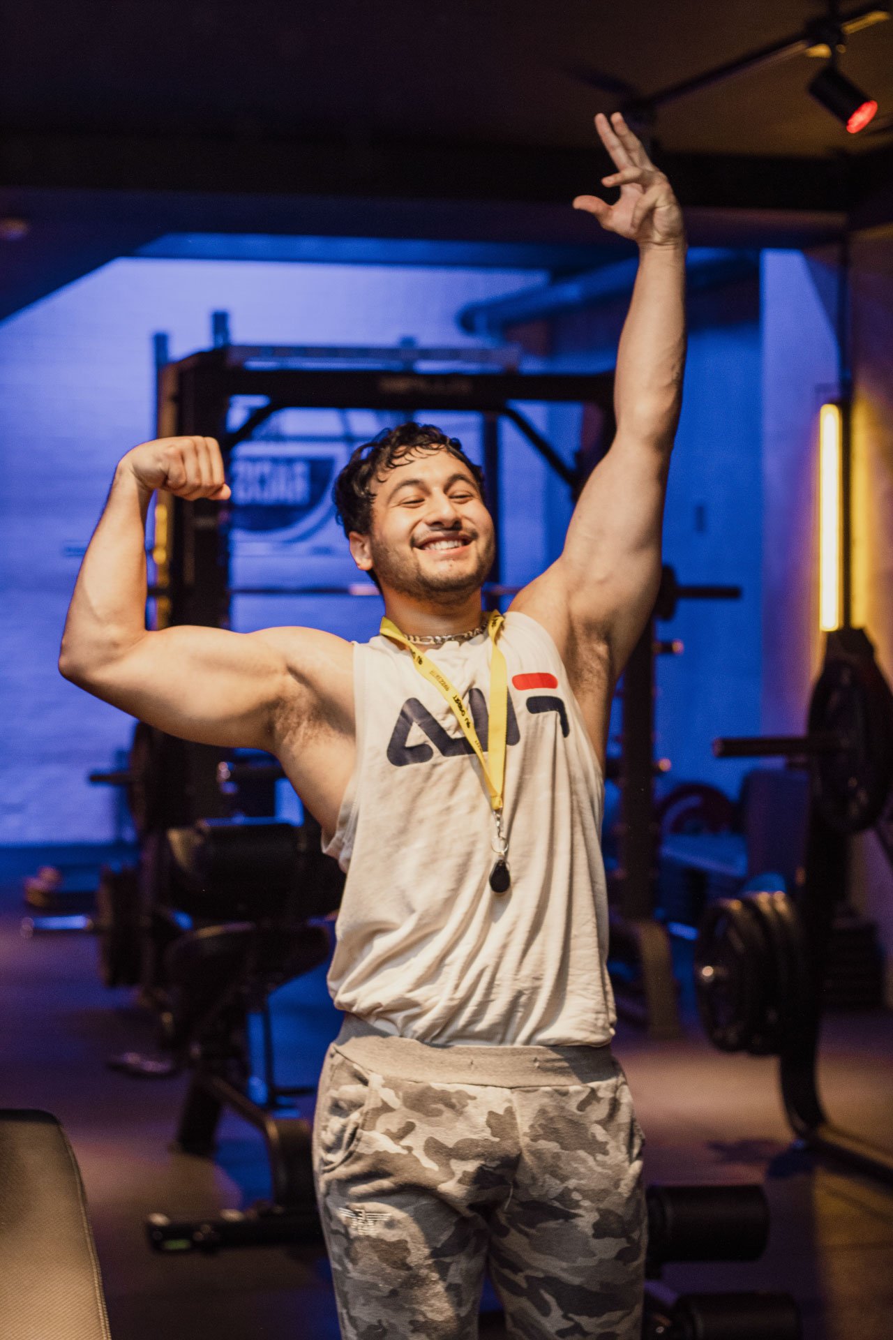 Homem sorridente posando no ginásio, usando uma camisa sem mangas, calça camuflada e uma medalha ao redor do pescoço, enquanto mostra o bíceps com uma expressão de orgulho.