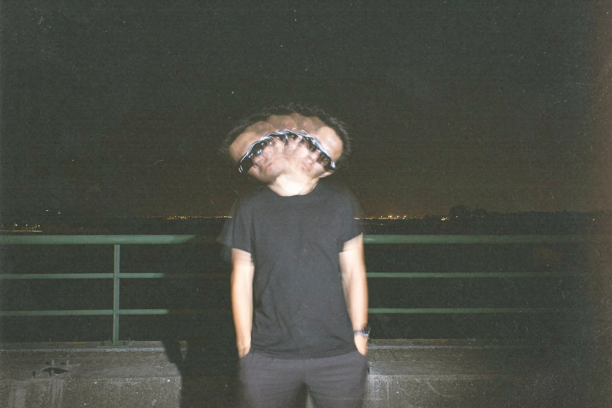 Pessoa com camisa preta e mãos nos bolsos, com efeito de distorção na cabeça, na noite, ao ar livre, com fundo de céu estrelado e horizonte distante com luzes urbanas.