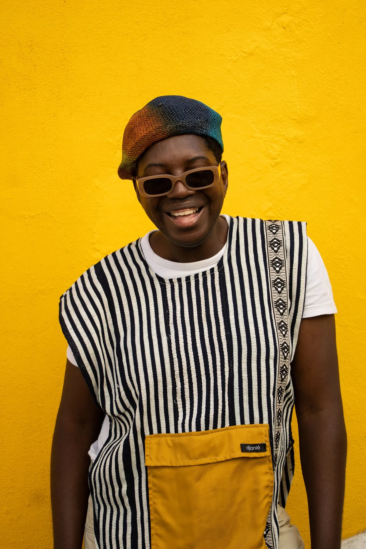 Pessoa sorrindo vestindo óculos escuros, chapéu colorido, camiseta de listras e avental amarelo em parede amarela.