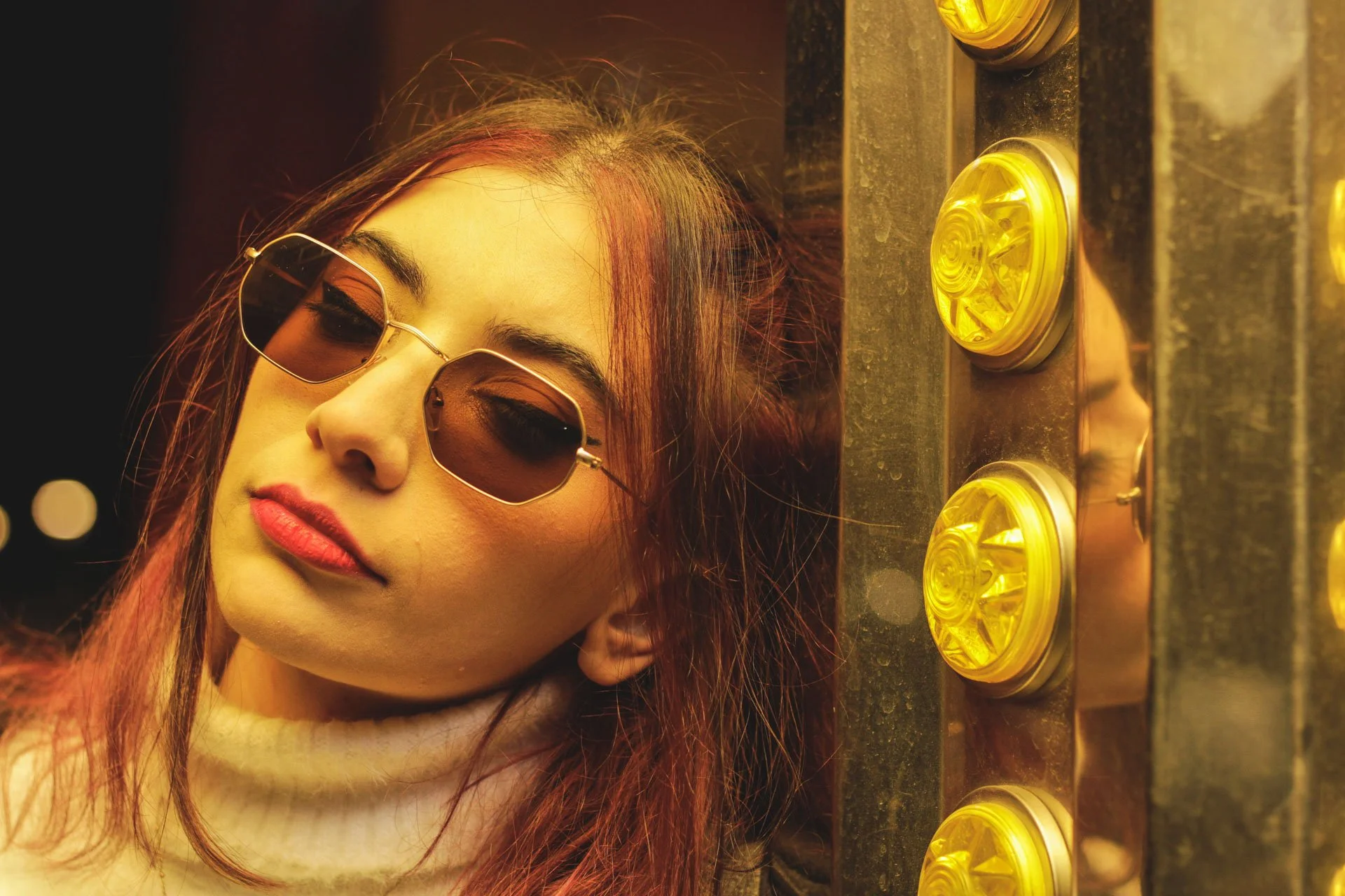 Mulher com cabelo vermelho e sombra nos olhos, usando óculos escuros, com roupas brancas, encostada em uma parede com luzes amarelas ao lado.