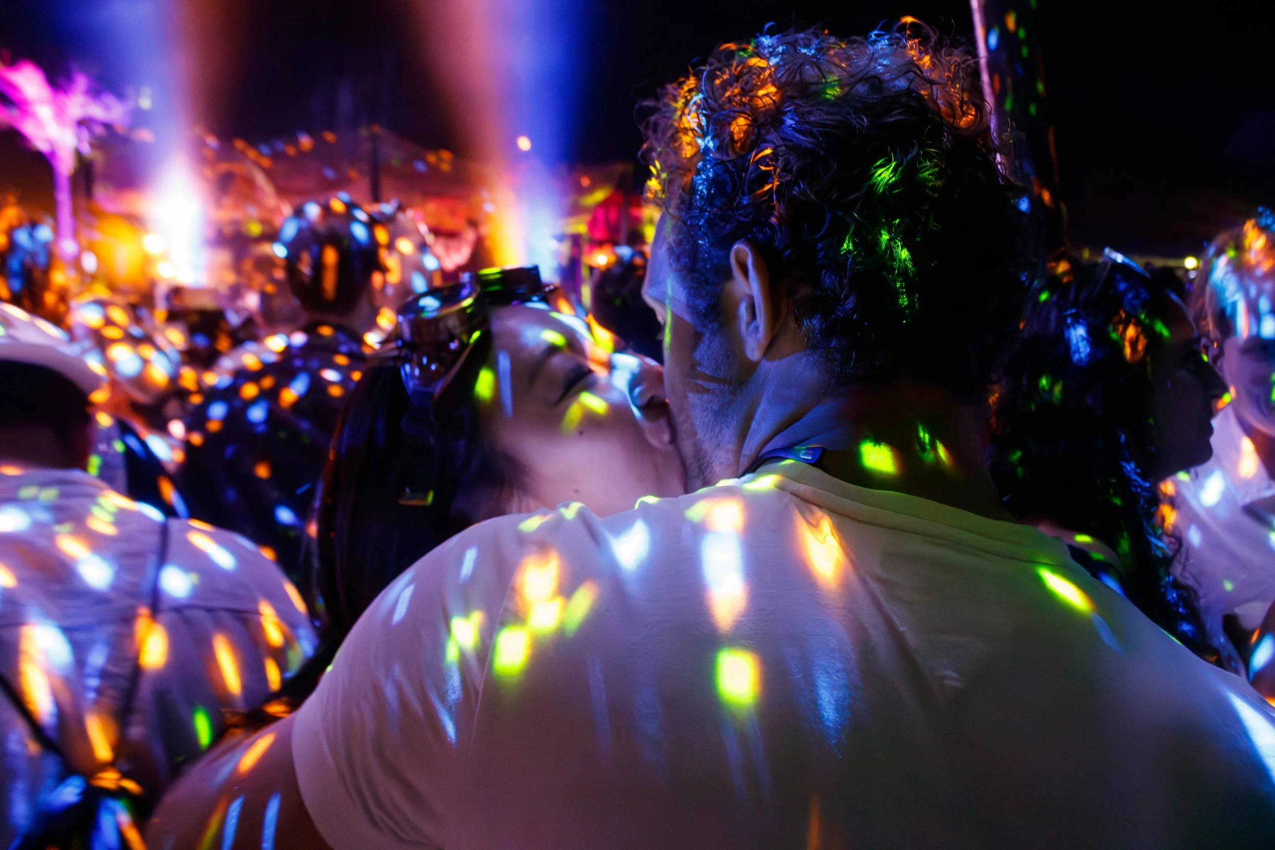 Casal se beijando em meio a uma multidão com luzes coloridas e efeito de luzes neon em uma festa ou evento noturno.