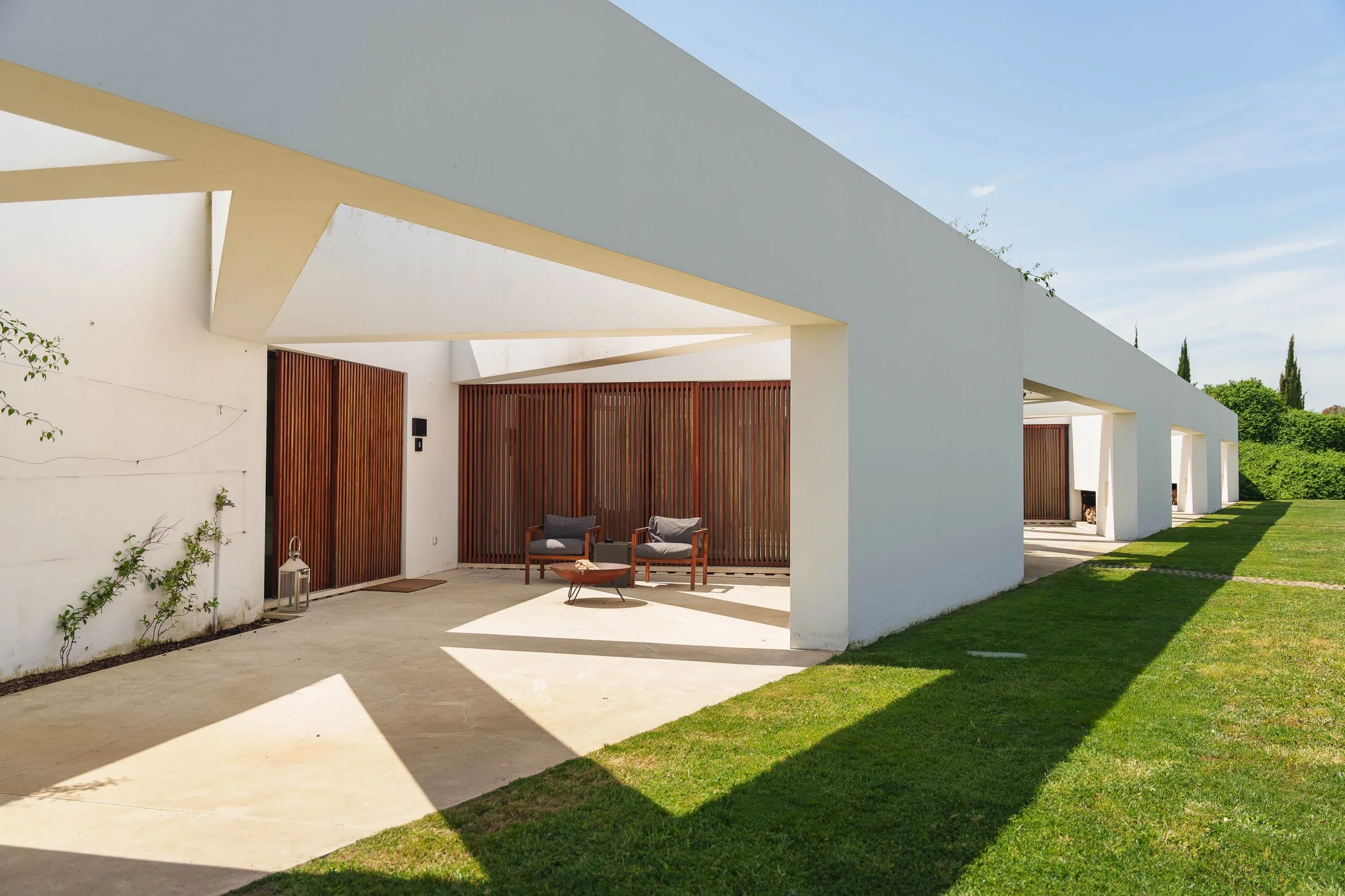 Projéteis brancos com aberturas em forma de retângulo, com paredes de ripas de madeira, jardim com grama e plantas verdes ao redor, céu azul ao fundo.