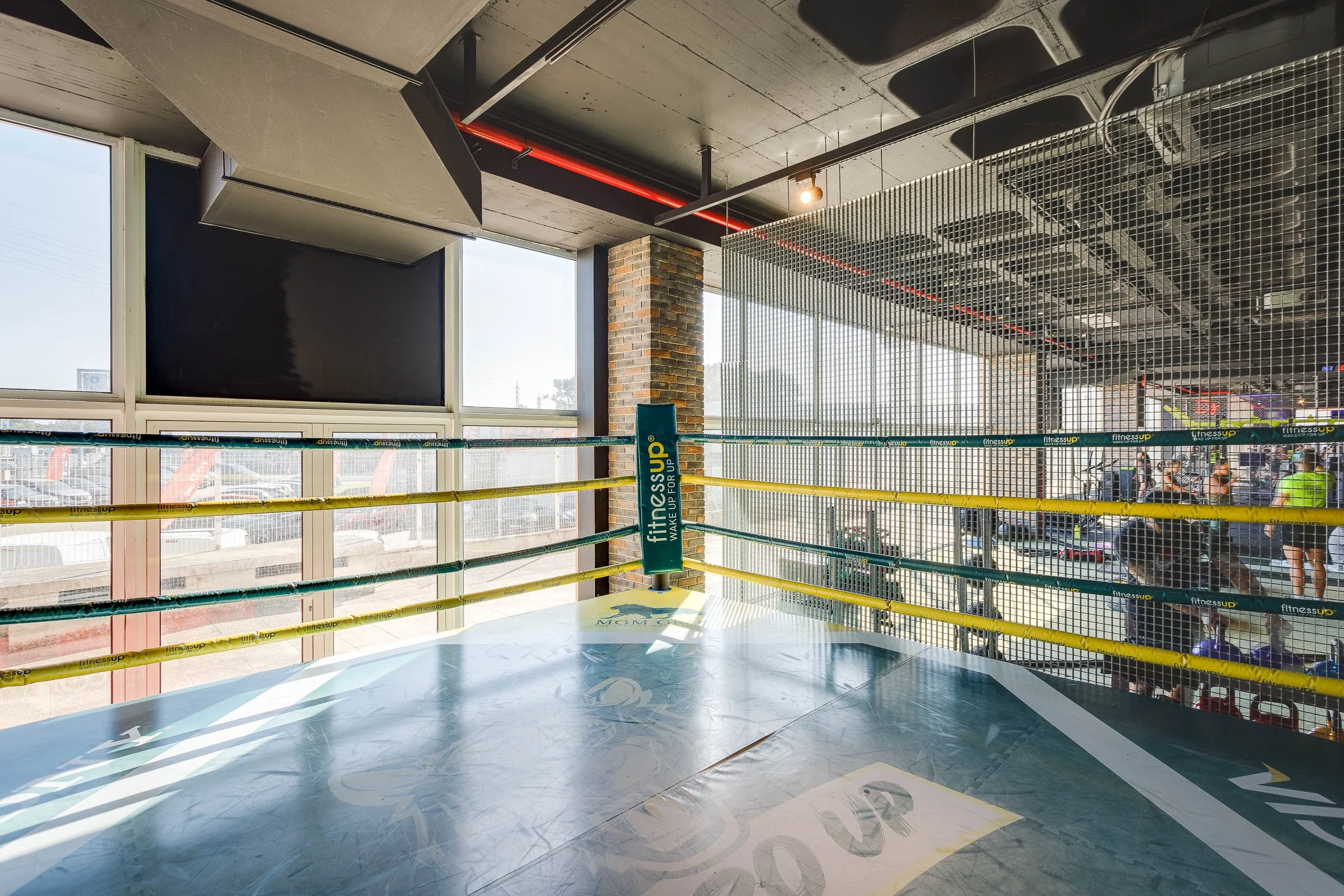 Ringue de boxe em uma academia, com iluminação natural, paredes de vidro e uma estrutura de aço.