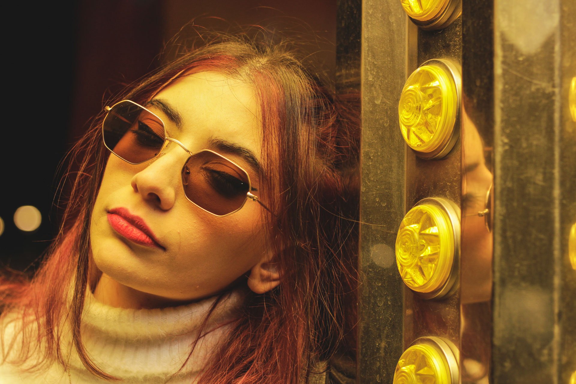 Jovem mulher com cabelo avermelhado, usando óculos de sol escuros e um suéter branco, apoiada na parede de uma cabine de foto com luzes amarelas.