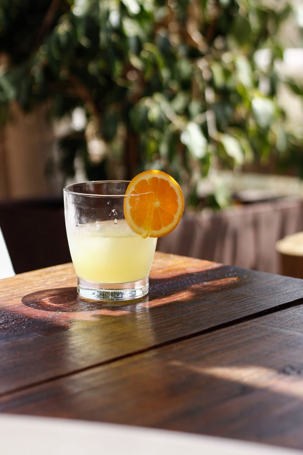 Copo de vidro com uma bebida amarelada e uma rodela de laranja na borda, colocado sobre uma mesa de madeira.