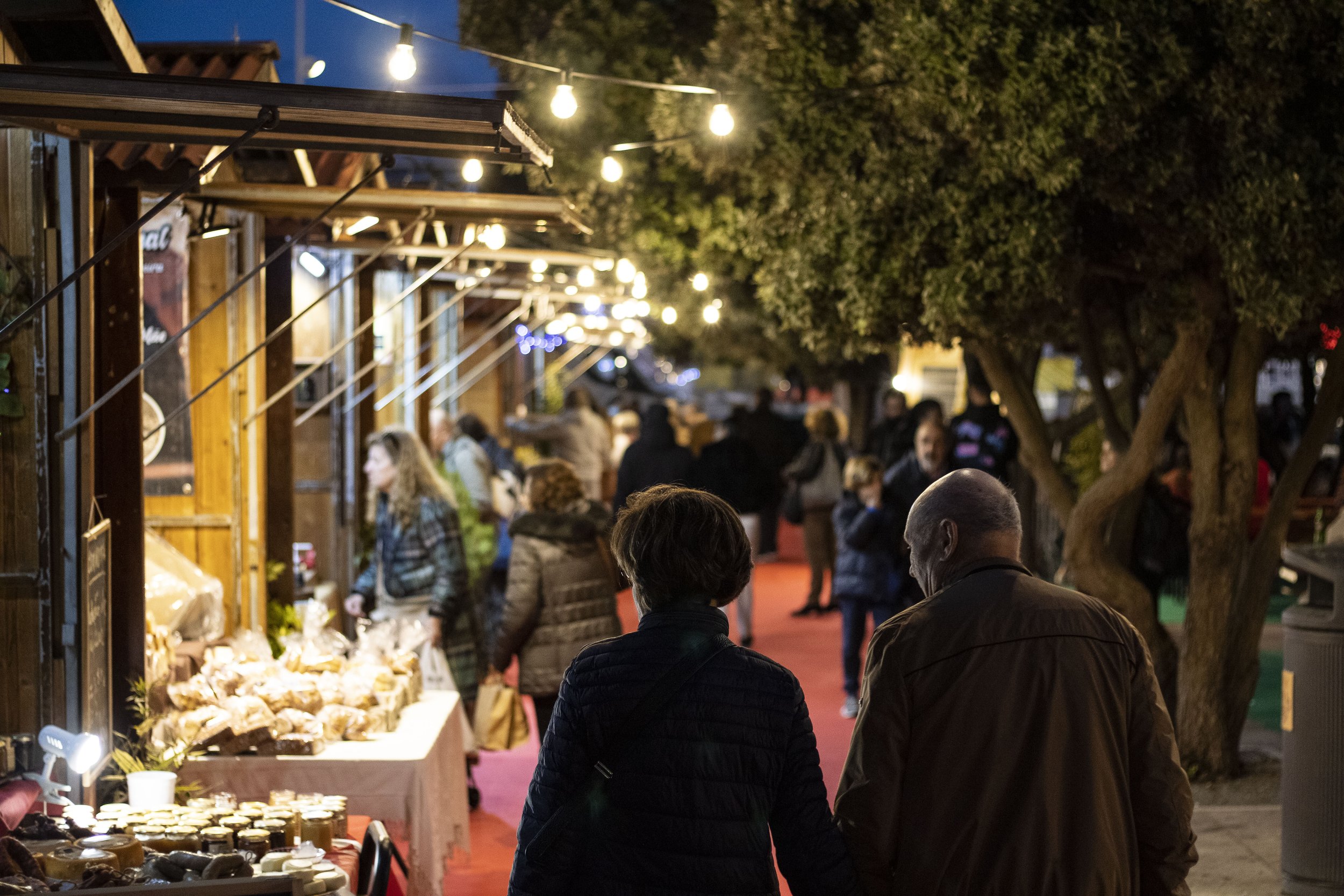 Casal de idosos caminhando de mãos dadas em feira ao ar livre à noite, com barracas de comida e outras pessoas ao fundo.