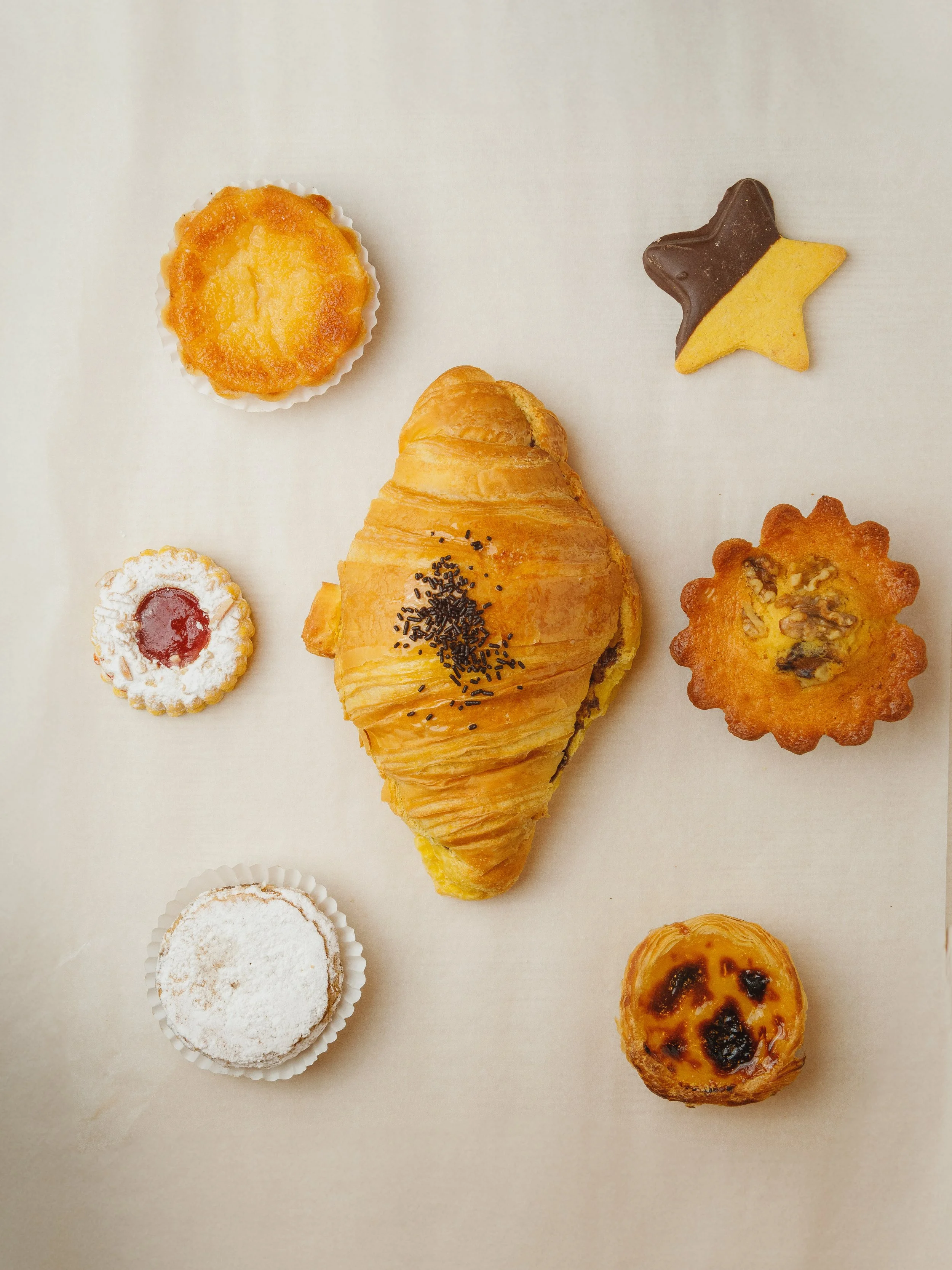 Vários tipos de doces e salgados, incluindo um croissant, muffins, cookies e um pão de queijo, dispostos sobre uma superfície clara.