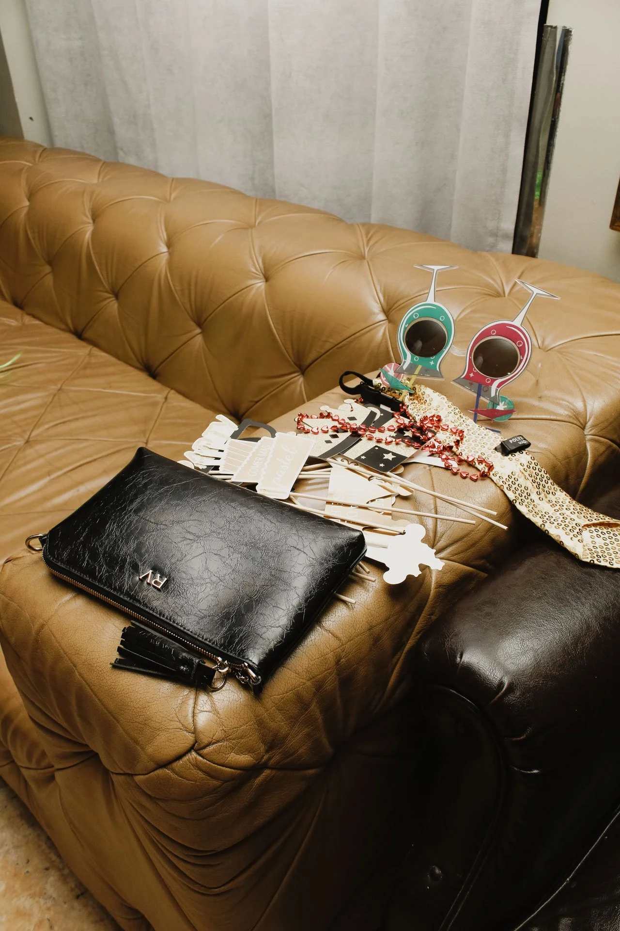 Sofá de couro em tom marrom com itens de festa espalhados, incluindo óculos de papel decorativos, faixa de galinha, acessórios de festa, e uma bolsa preta.