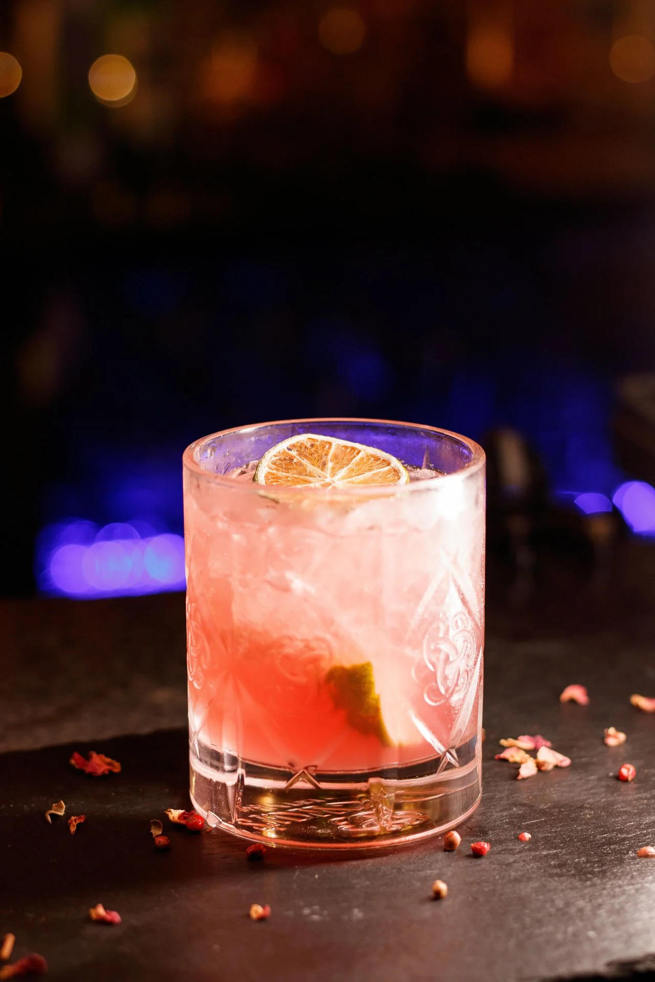 Copo de vidro com bebida rosa, gelo, uma fatia de limão na borda e uma fatia dentro, em uma mesa com sementes de pimenta espalhadas ao redor, fundo escuro com luzes difusas