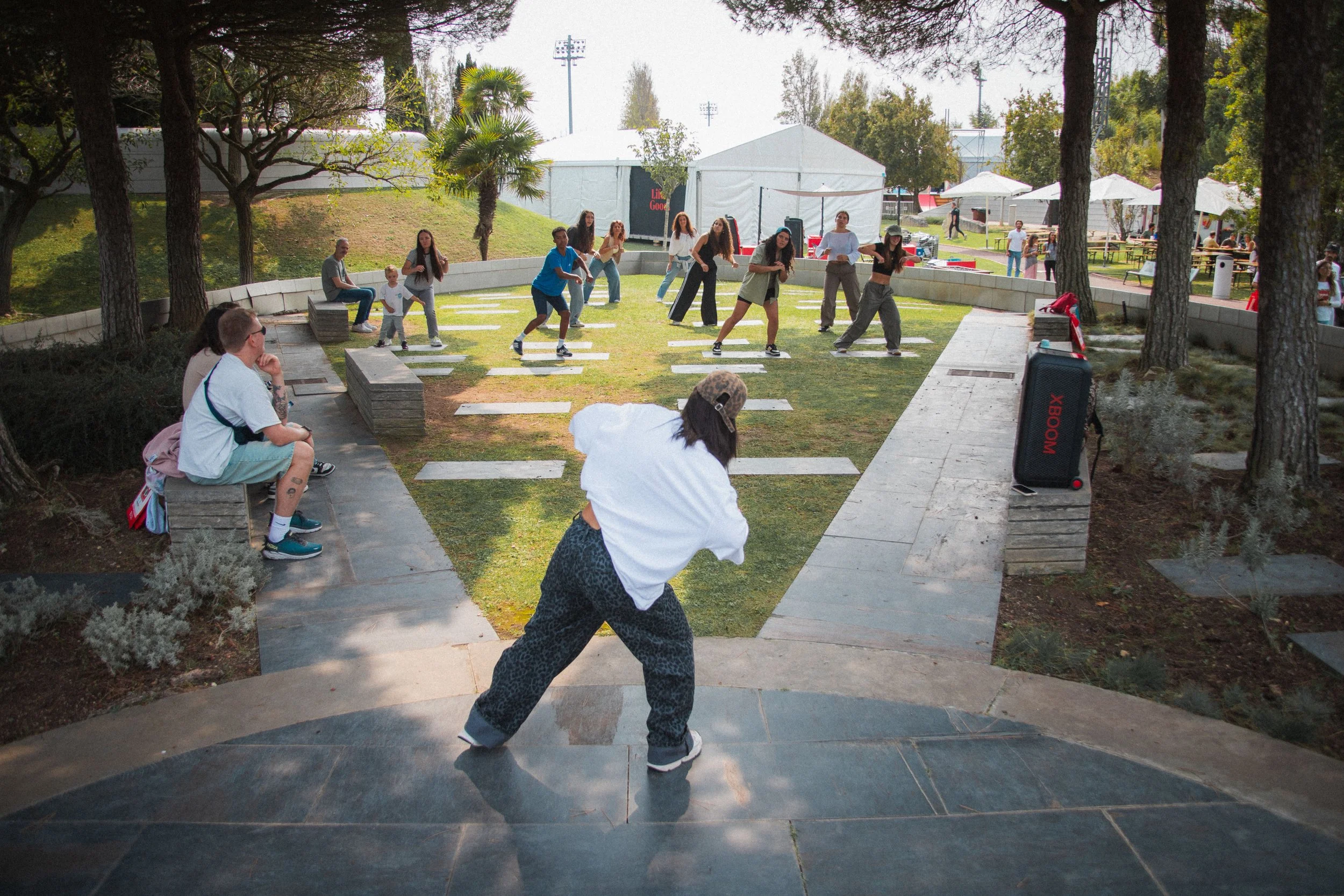 Grupo de pessoas assistindo uma aula de dança ao ar livre em um parque com árvores, gramado e iluminação artificial ao fundo.