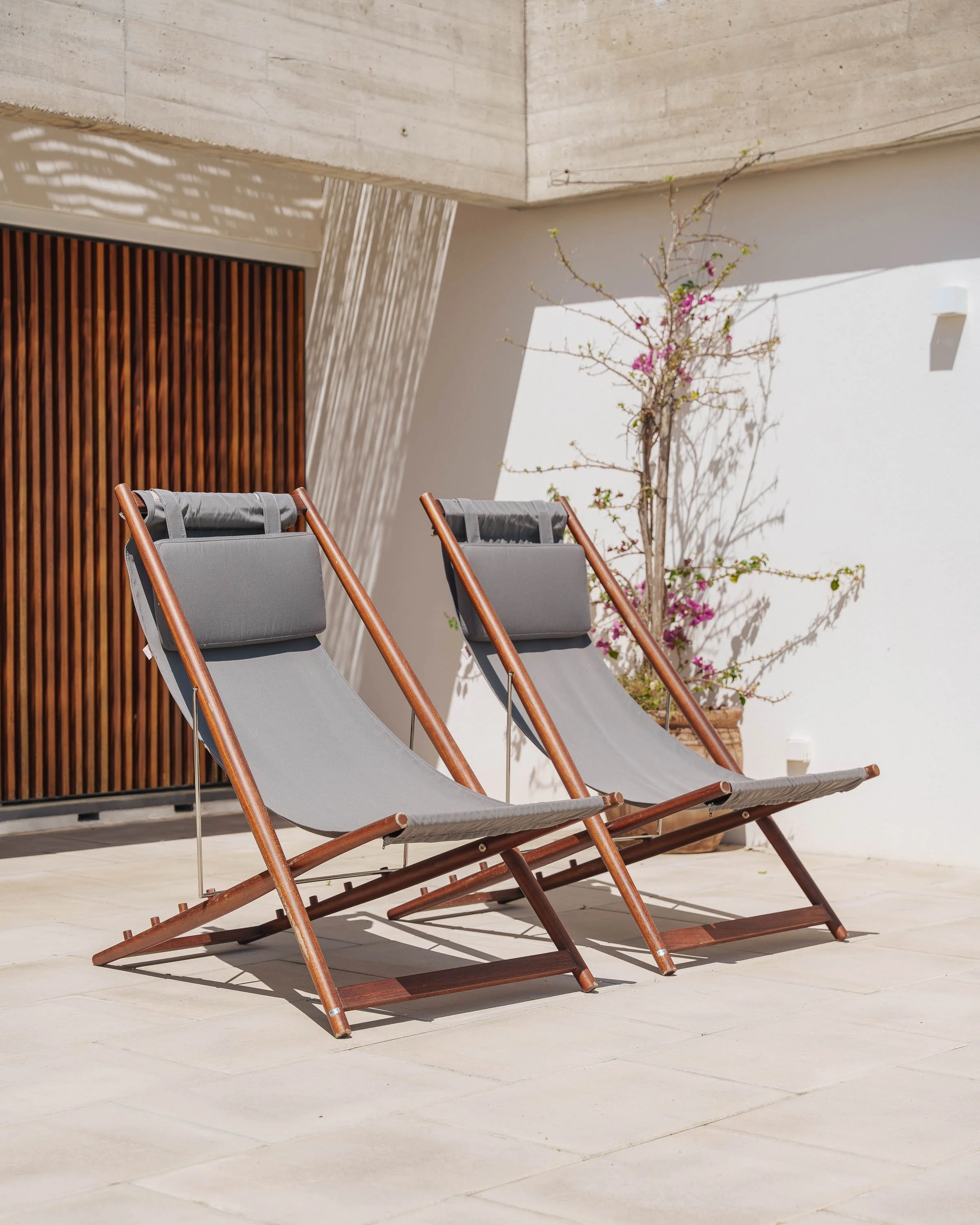 Duas cadeiras de descanso de madeira com almofadas cinzas, colocadas ao lado de uma planta com flores rosas, em um espaço ao ar livre com piso de pedra e parede branca.