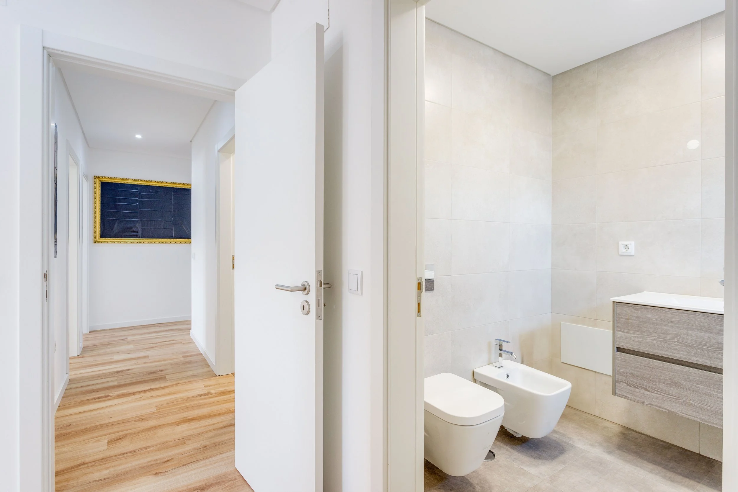 Corredor com portas para diferentes cômodos e uma parede decorada com moldura dourada, incluindo um banheiro com vaso sanitário, bidê, pia de madeira e parede revestida de azulejos bege.