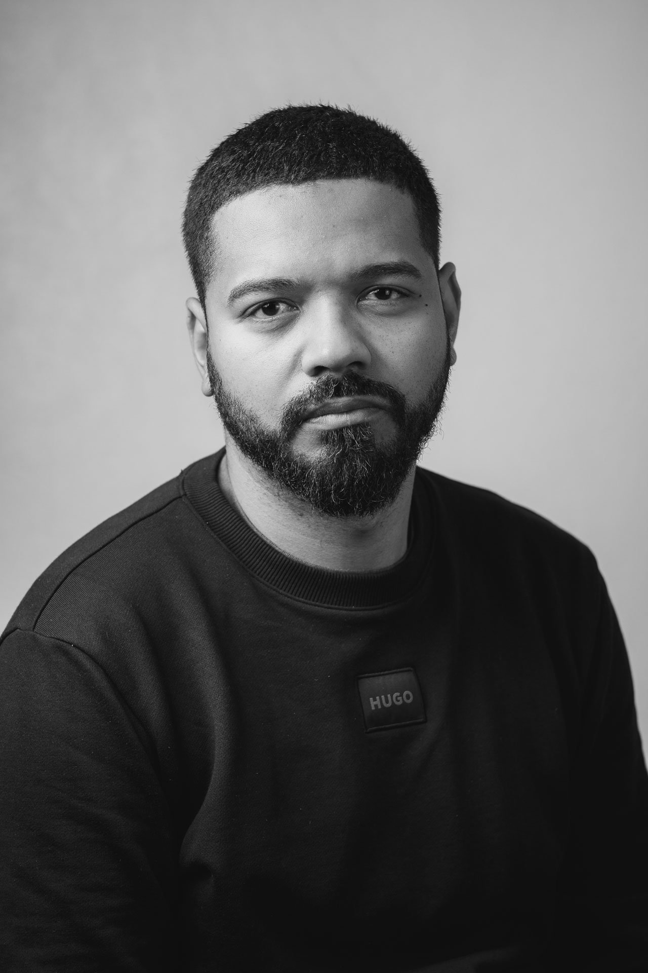 Retrato de um homem com cabelo curto, barba por fazer, usando uma blusa preta com um rótulo que diz "HUGO" na frente, em uma fotografia em preto e branco com fundo neutro.