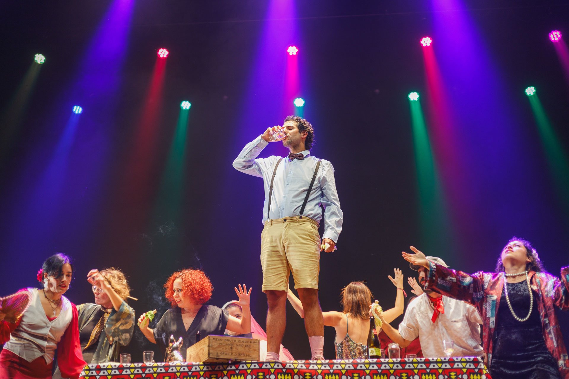 Homem com roupas vintage, de shorts e suspensórios, tomando uma bebida no palco com várias pessoas ao redor durante uma apresentação teatral ou musical com iluminação colorida.