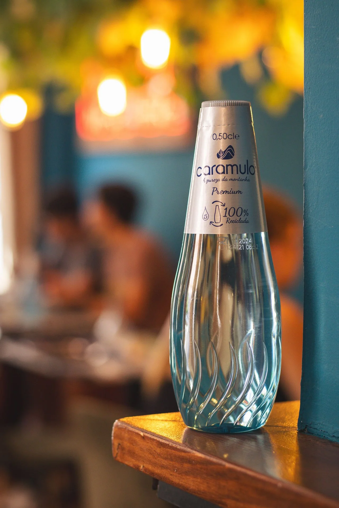 Garrafa de água de 0,5L, transparente, com rótulo de papel prateado com informações de marca e reciclagem, apoiada em mesa de madeira em ambiente com luzes amareladas e pessoas ao fundo.