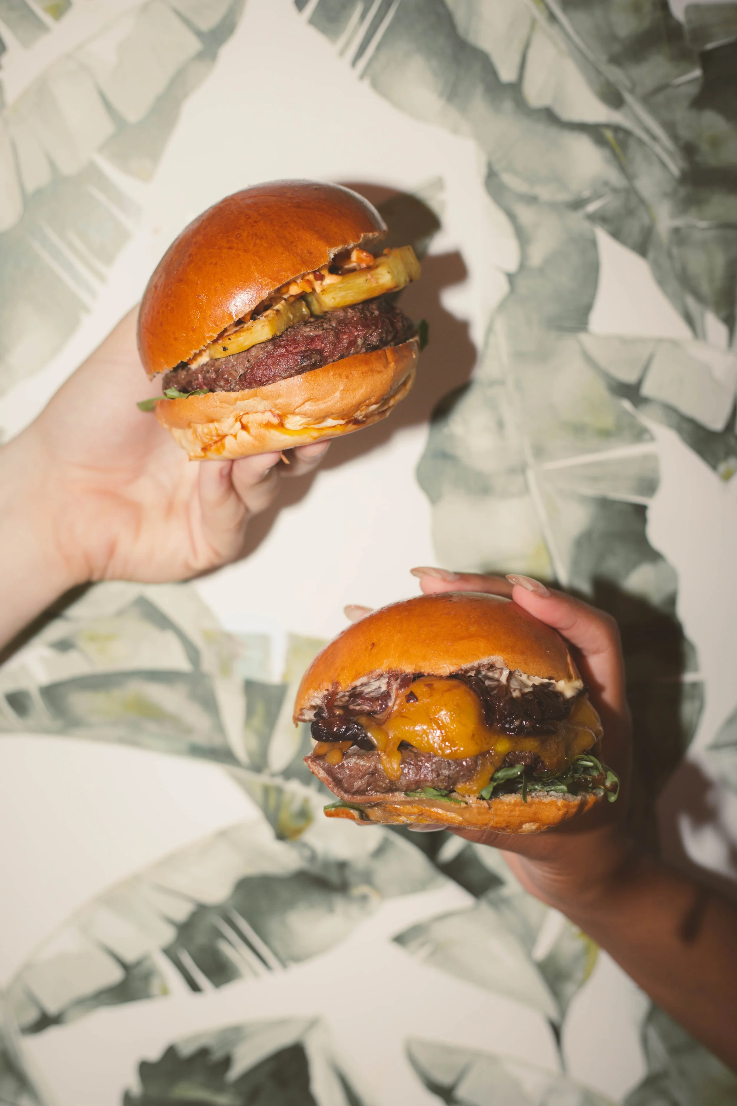 Duas mãos segurando hambúrgueres com carne, queijo, molho e outros ingredientes em pães dourados, sobre uma superfície com papel de parede com estampa de folhas verdes.