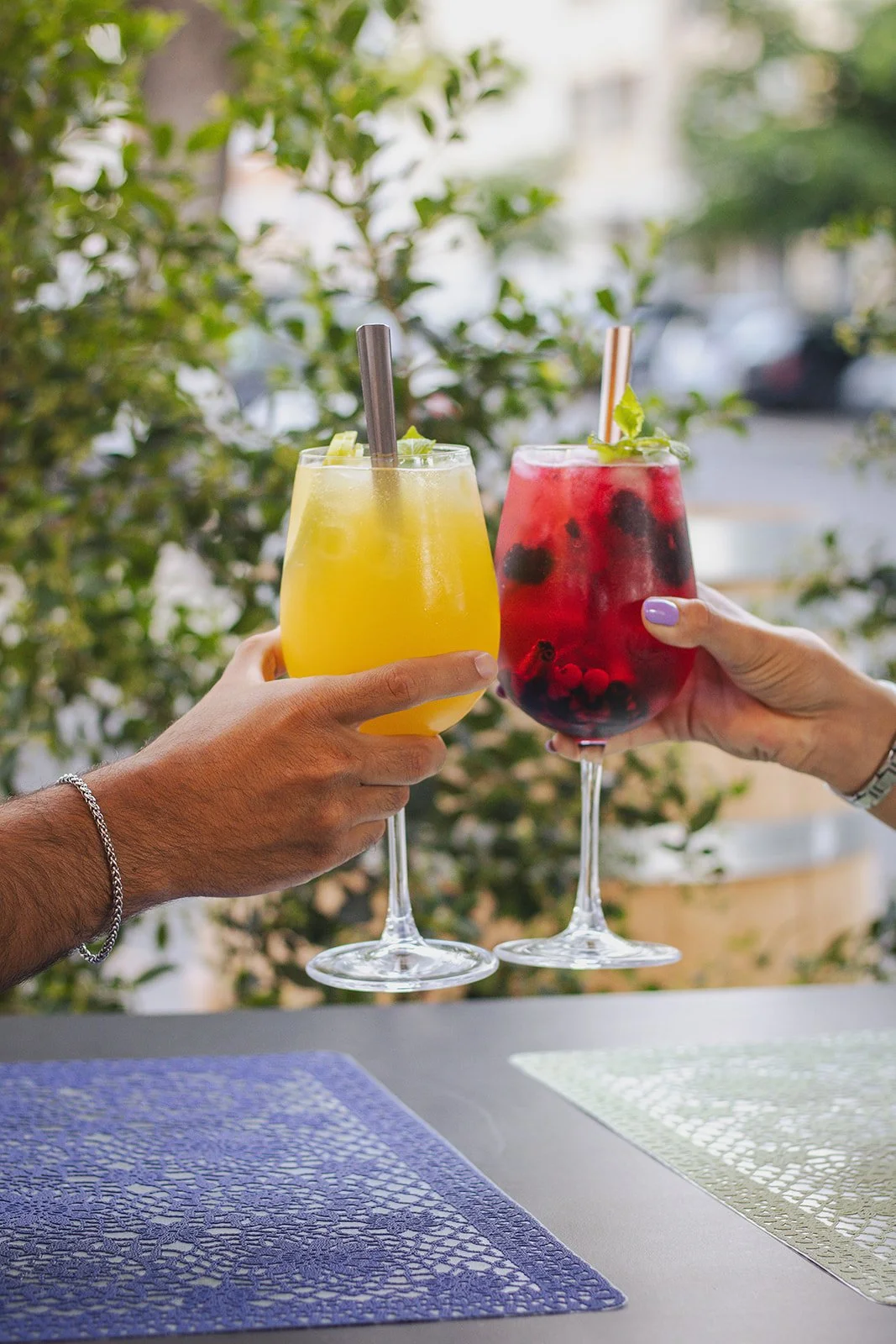 Duas mãos segurando taças de drinks coloridos, um de marrom e outro de vermelho, com frutas e hortelã, contra fundo de árvores e carros na rua.