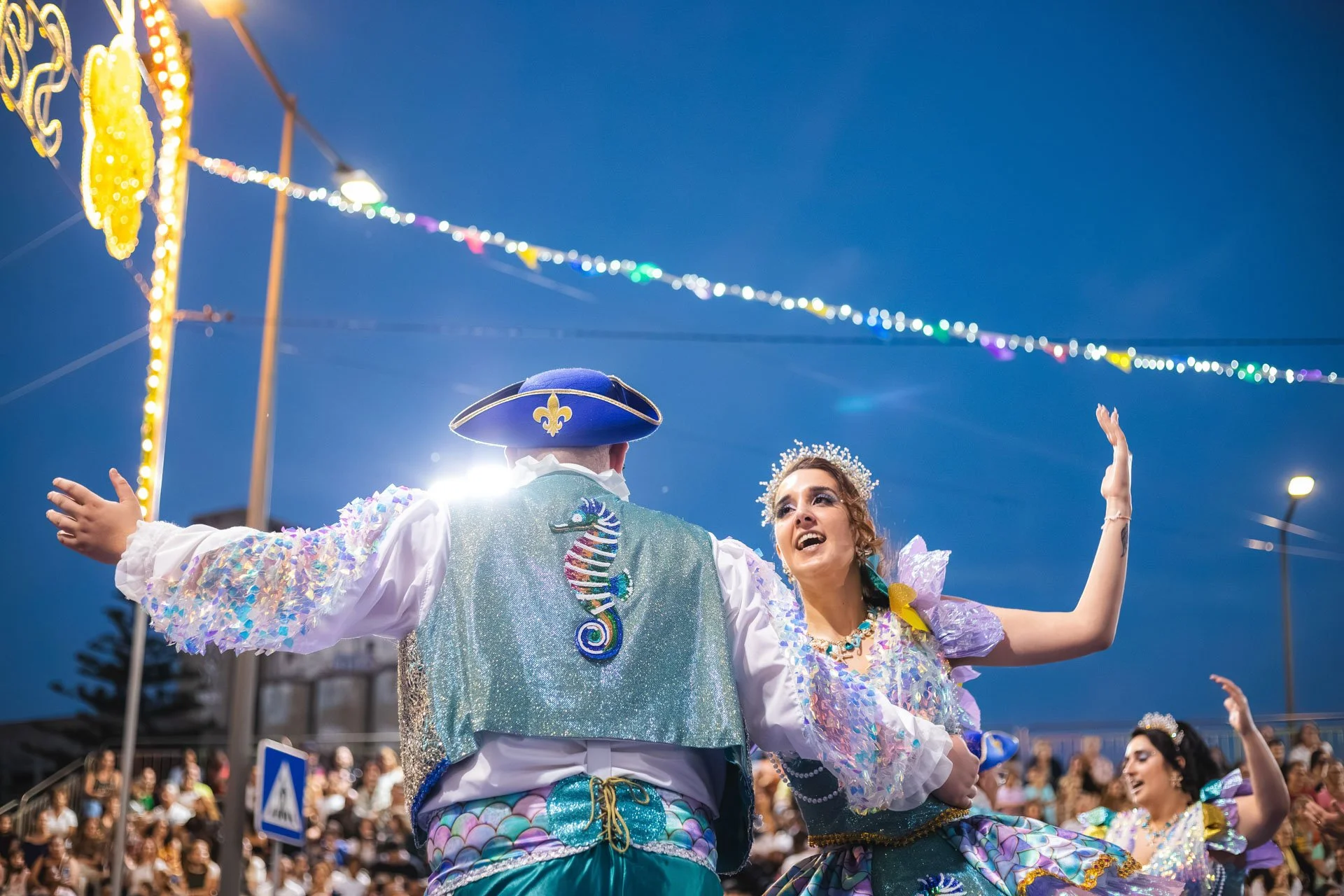 Duas pessoas vestidas com fantasias brilhantes e coloridas fazendo uma dança durante uma celebração ao ar livre à noite, com uma multidão assistindo ao fundo e luzes decorativas no céu.