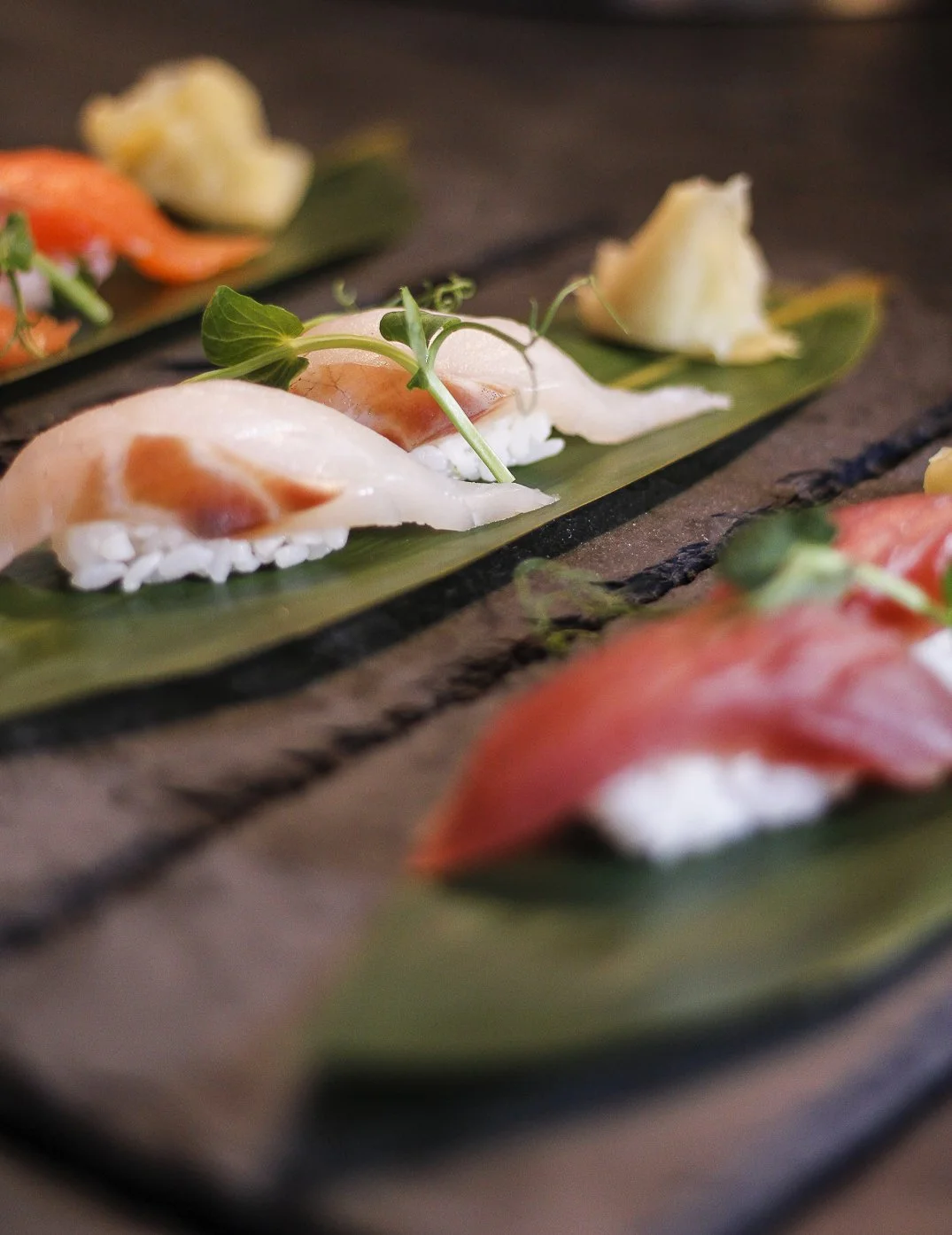 Sushi de peixe branco com microverdes, servido sobre folha de bananeira, fundo desfocado.