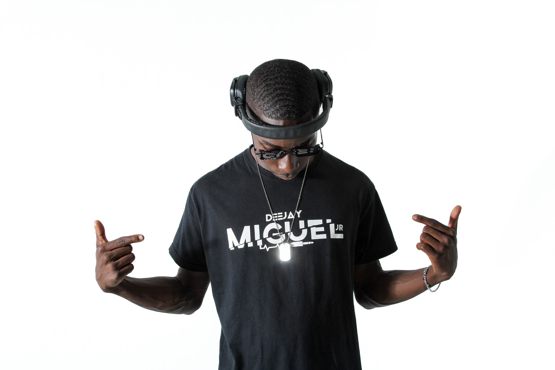 Homem negro usando fones de ouvido pretos, óculos escuros e camiseta preta com o nome 'MIGUEL' em destaque, apontando para si mesmo com os dedos, contra fundo branco.