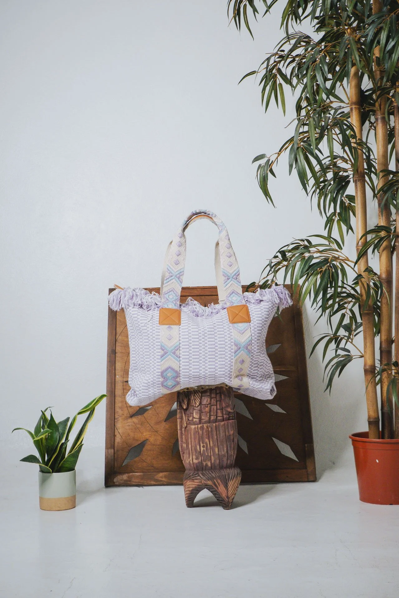 Bolsa de ombro branca com detalhes em padrão pastel, apoiada em uma escultura de madeira, ao lado de plantas verdes, uma delas em vaso branco e a outra em vaso laranja, e uma placa de madeira ao fundo.