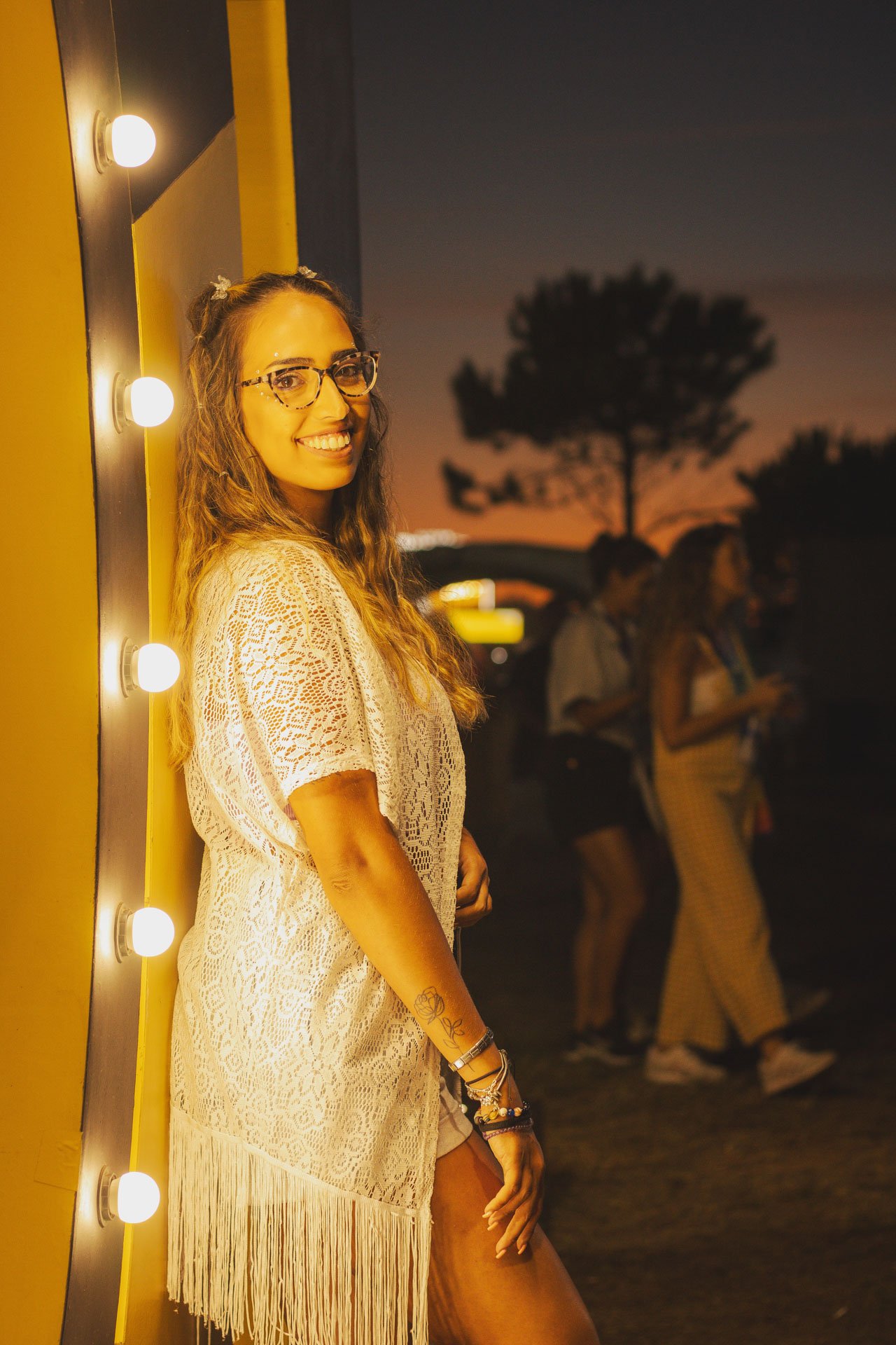Mulher sorridente com óculos de armação tartaruga, vestindo uma blusa de renda branca com franjas na ponta, de cabelo castanho claro, em uma rua ao pôr do sol, ao lado de uma parede com luzes de lâmpada.