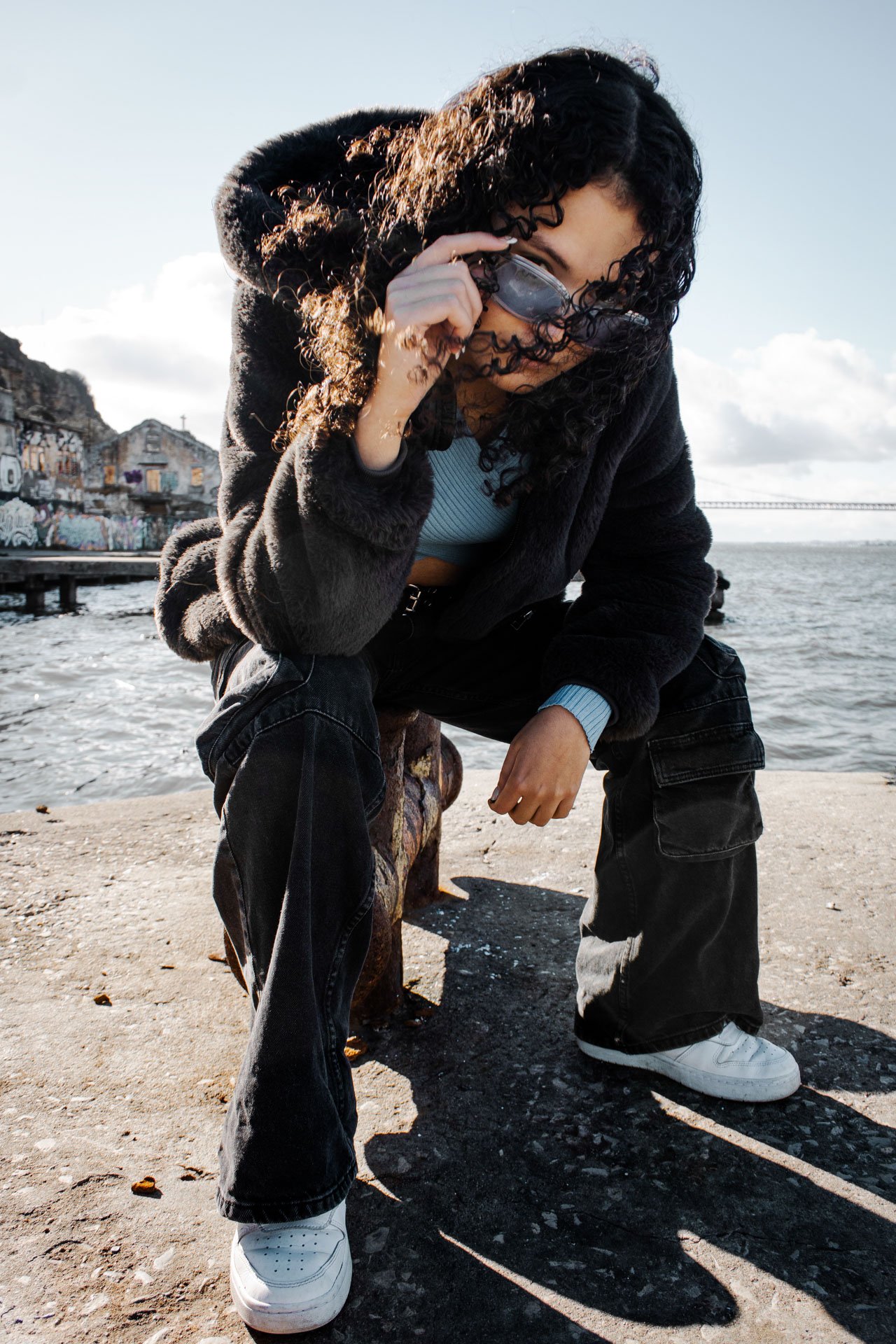 Jovem com cabelos cacheados escuros usando jaqueta preta, óculos de proteção e calça larga, posando perto do mar em uma rua de bairro