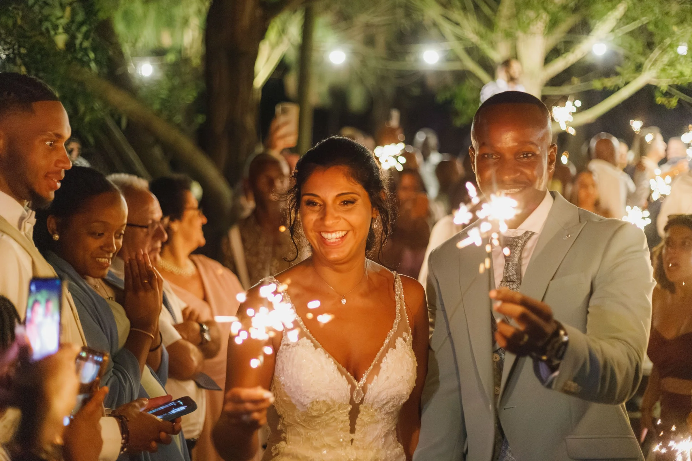 Casal sorridente em festa de casamento, segurando fogo de artifício.