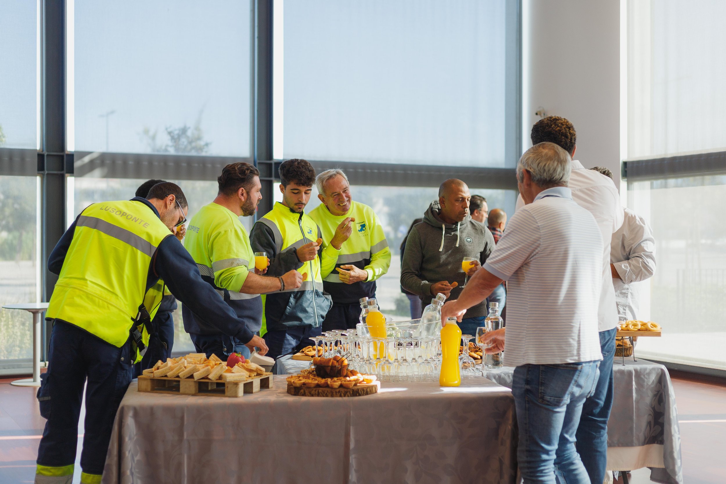 Pessoas em uma reunião de café da manhã em uma sala com janelas grandes, alguns vestindo jaquetas de serviço de emergência, servindo e desfrutando de alimentos e bebidas em uma mesa decorada com sucos, doces e sanduíches.