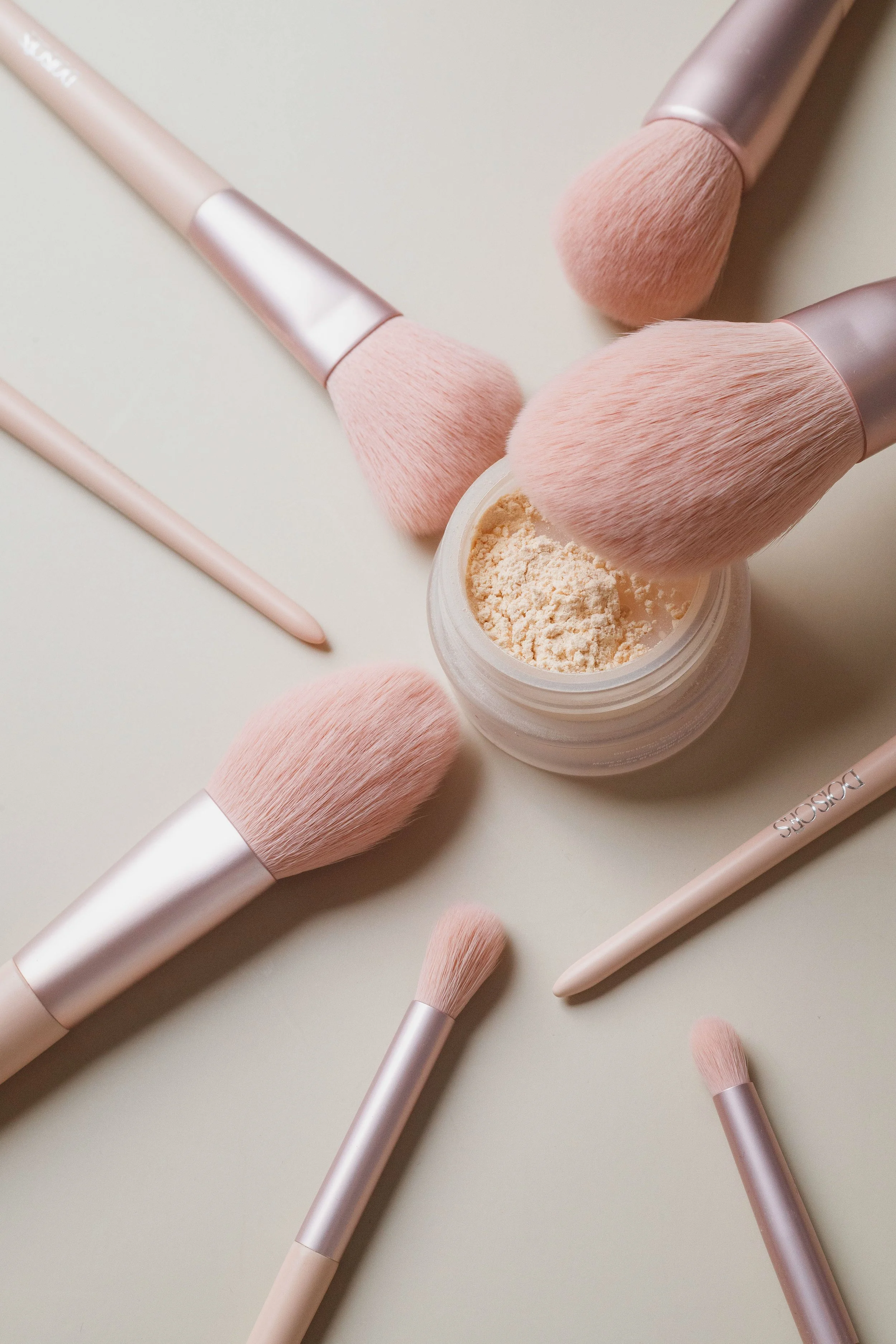Pincéis de maquiagem de cabo rosé, alguns com cerdas rosé e outros com pó compacto em pote branco, em fundo claro.